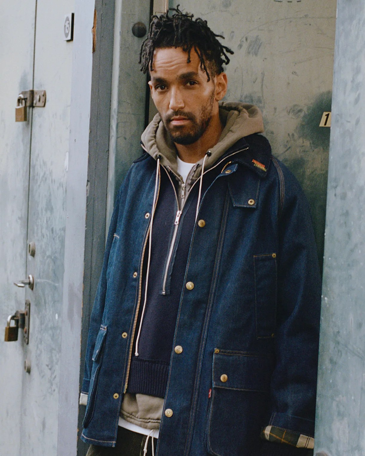 Man wearing layered clothing including a dark blue denim jacket with brass buttons and a beige hooded sweatshirt.
