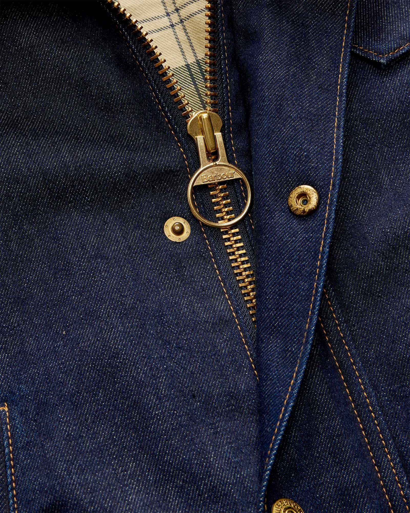 Close-up of a dark blue denim jacket with a gold zipper pull engraved with "Barbour" and brass snap buttons.