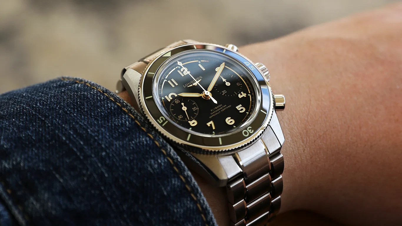 Silver metal chronograph wristwatch with black dial and gold numerals worn on a wrist next to a denim jacket sleeve.