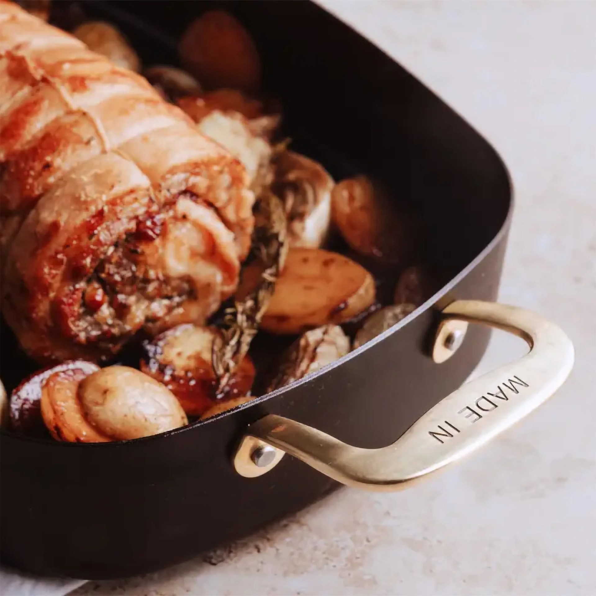 Close-up of a black roasting pan with brass handles labeled "MADE IN" containing roasted meat, mushrooms, and potatoes.