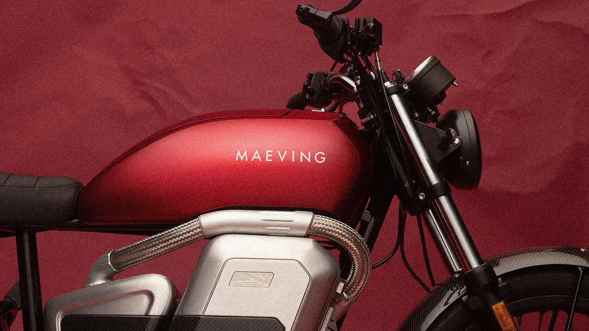 Red Maeving motorcycle fuel tank with silver braided hose and black handlebars against a red background.