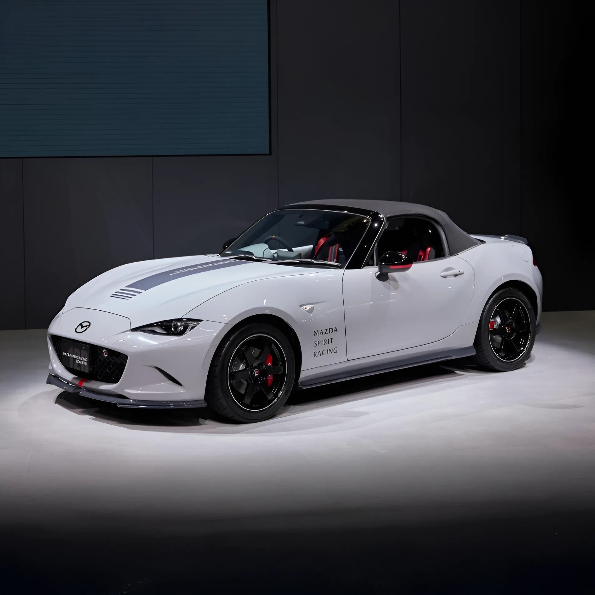 White Mazda convertible sports car with black soft top, black wheels, and "Mazda Spirit Racing" text on the door.