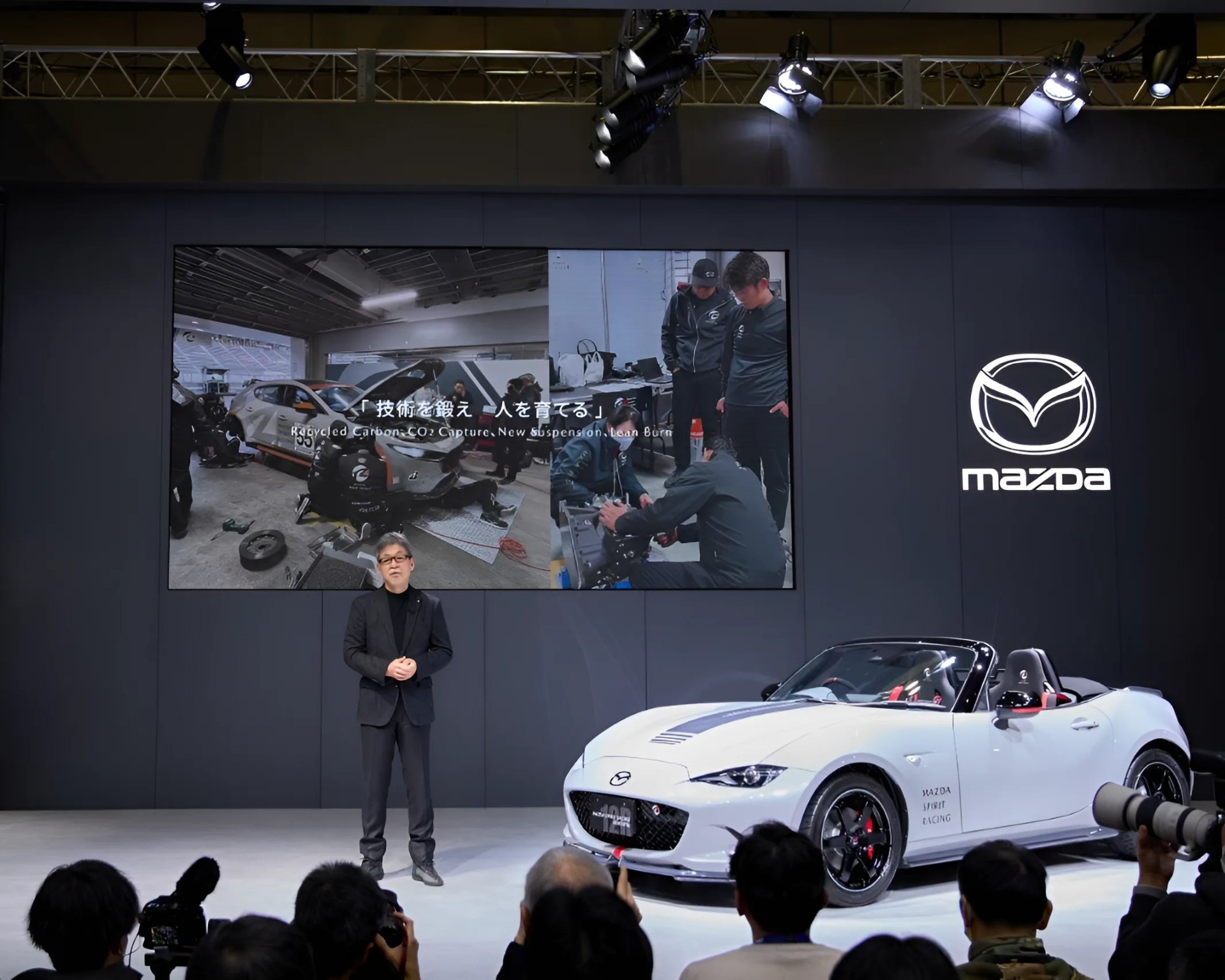 White Mazda convertible sports car with black and red accents displayed on stage next to a presenter and a large screen showing a workshop scene.