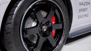 Glossy black five-spoke car wheel with red brake caliper and Yokohama tire on a white Mazda Spirit Racing car.