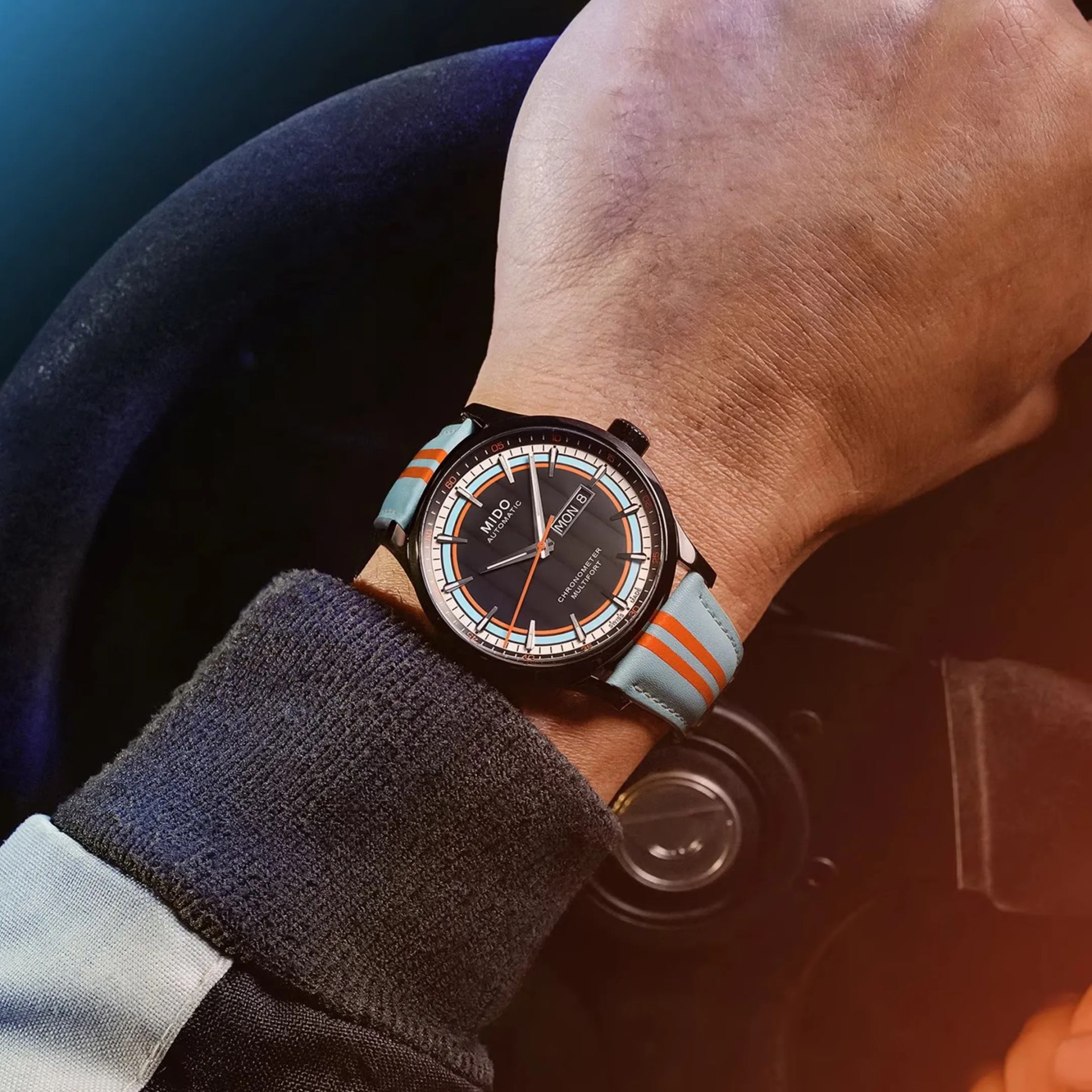 Black dial Mido Multifort chronometer watch with day-date display and light blue strap with orange stripes on a wrist.