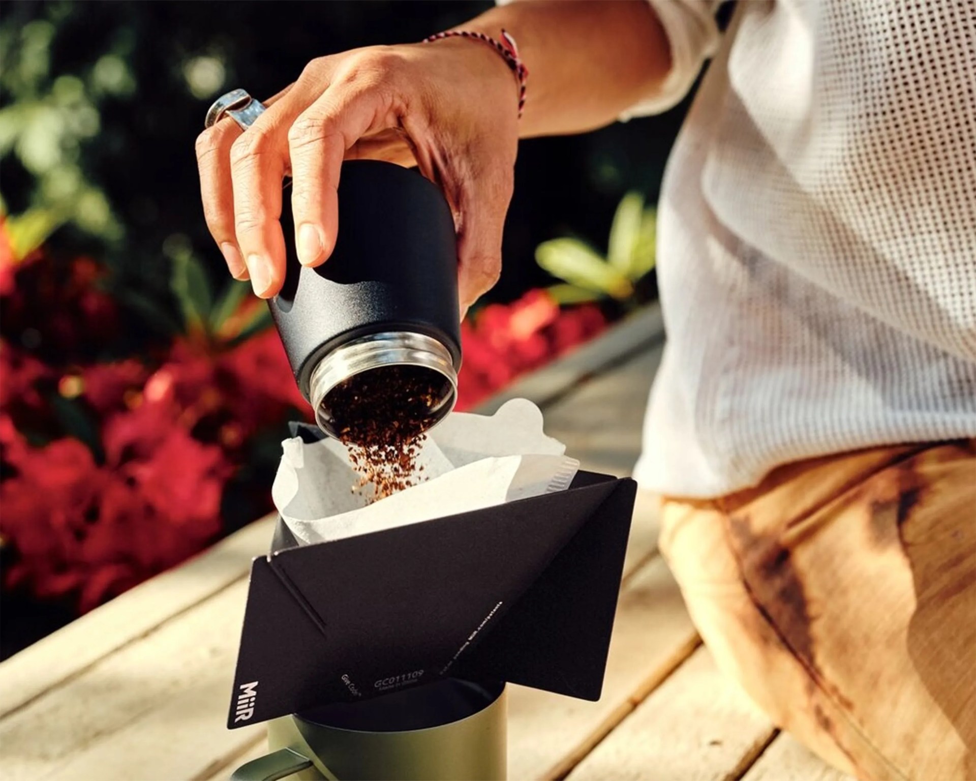 Hand pouring ground coffee from a black container into a white paper filter held by a black Miir coffee dripper over a green mug.