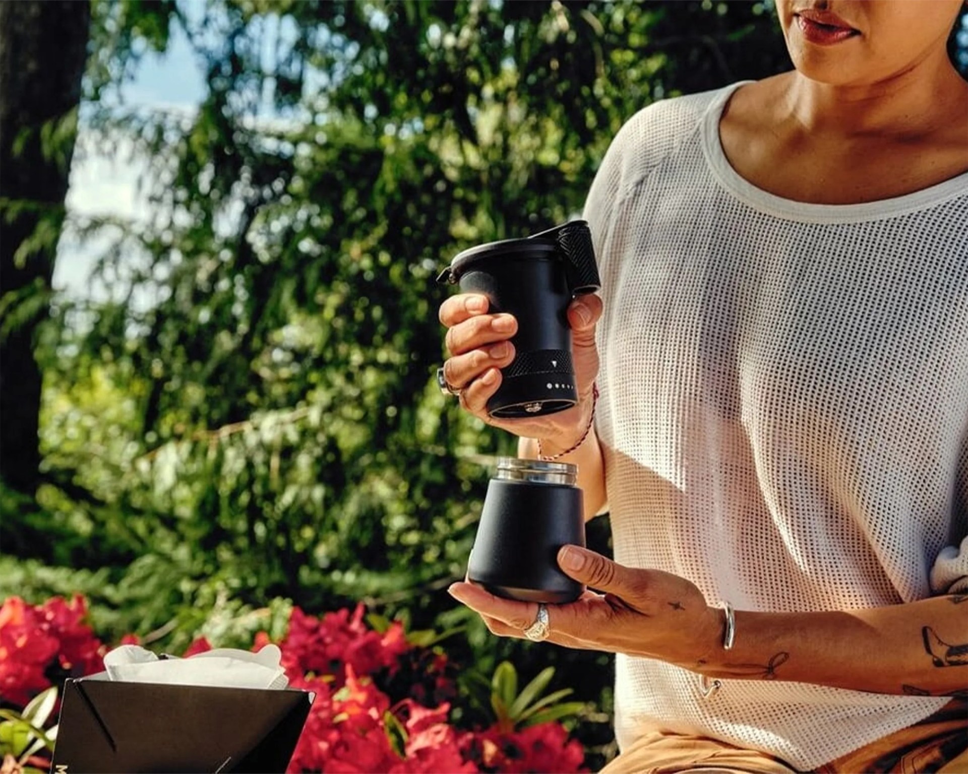 Person holding a black portable coffee maker with a detachable cup outdoors near red flowers and green trees.