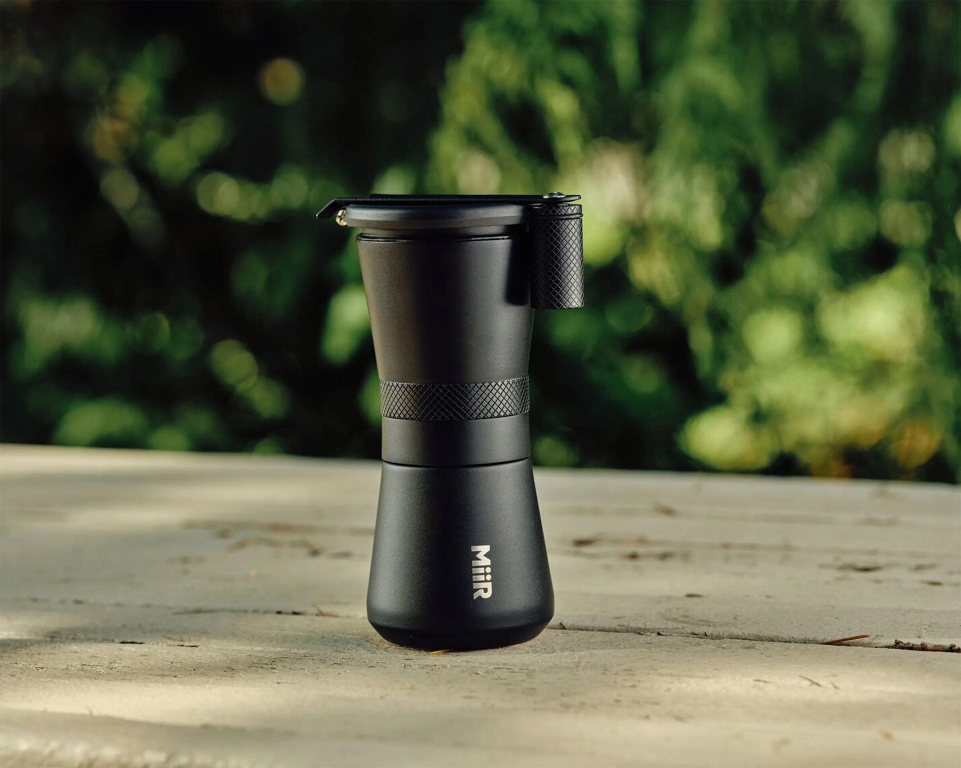 Black Miir insulated travel mug with textured grip and flip-top lid on an outdoor table.