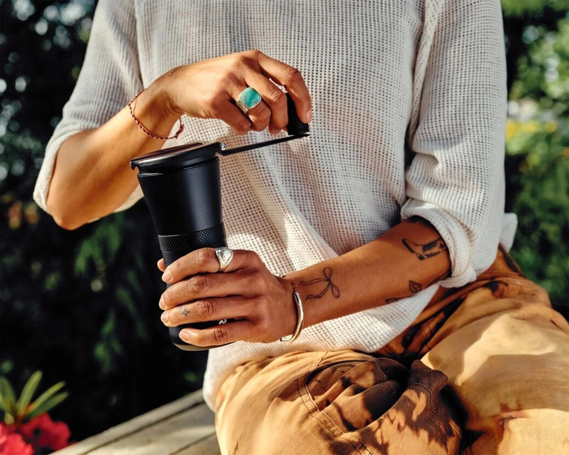 Person wearing a white knit sweater and tan pants holding a black manual coffee grinder outdoors.