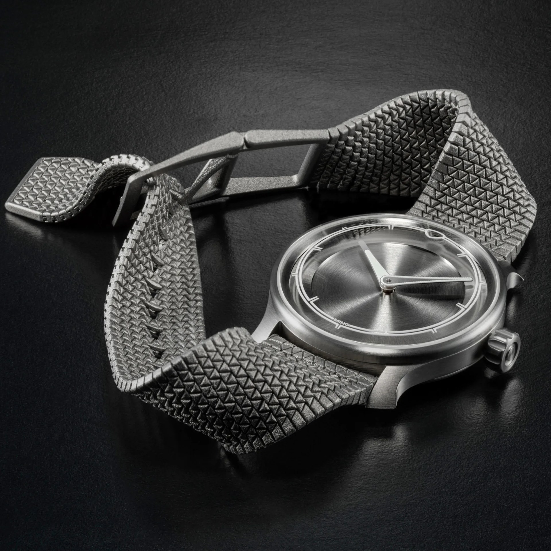 Silver wristwatch with a textured gray strap and a minimalist brushed metal dial on a black surface.