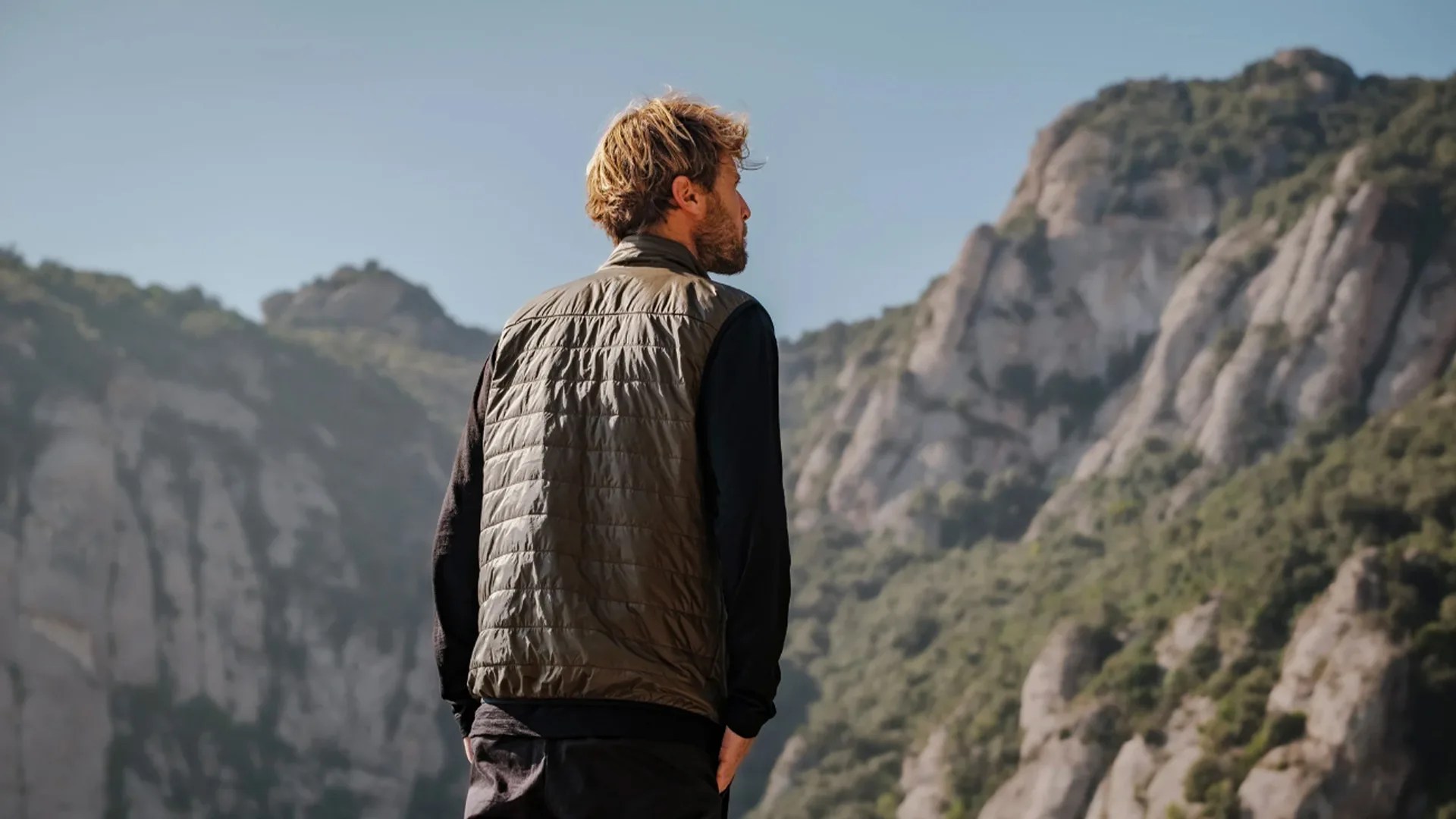Man wearing an olive green quilted vest over a black long-sleeve shirt standing outdoors with rocky mountains in the background.
