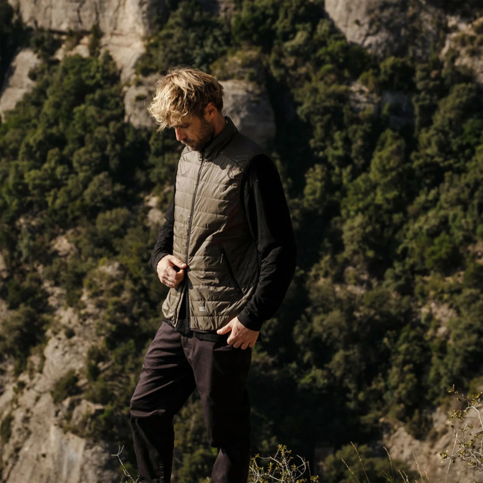 Man wearing an olive-green quilted vest over a black long-sleeve shirt standing outdoors with rocky cliffs and trees in the background.
