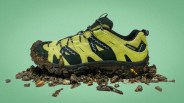 Yellow and black Gramicci trail shoe with mud on the sole and surrounding it on a green background.