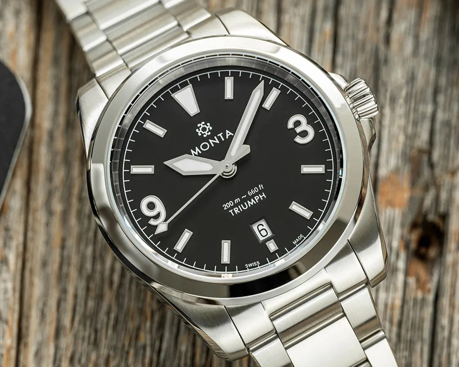 Stainless steel Monta Triumph wristwatch with black dial, white markers, and date window on wooden surface.