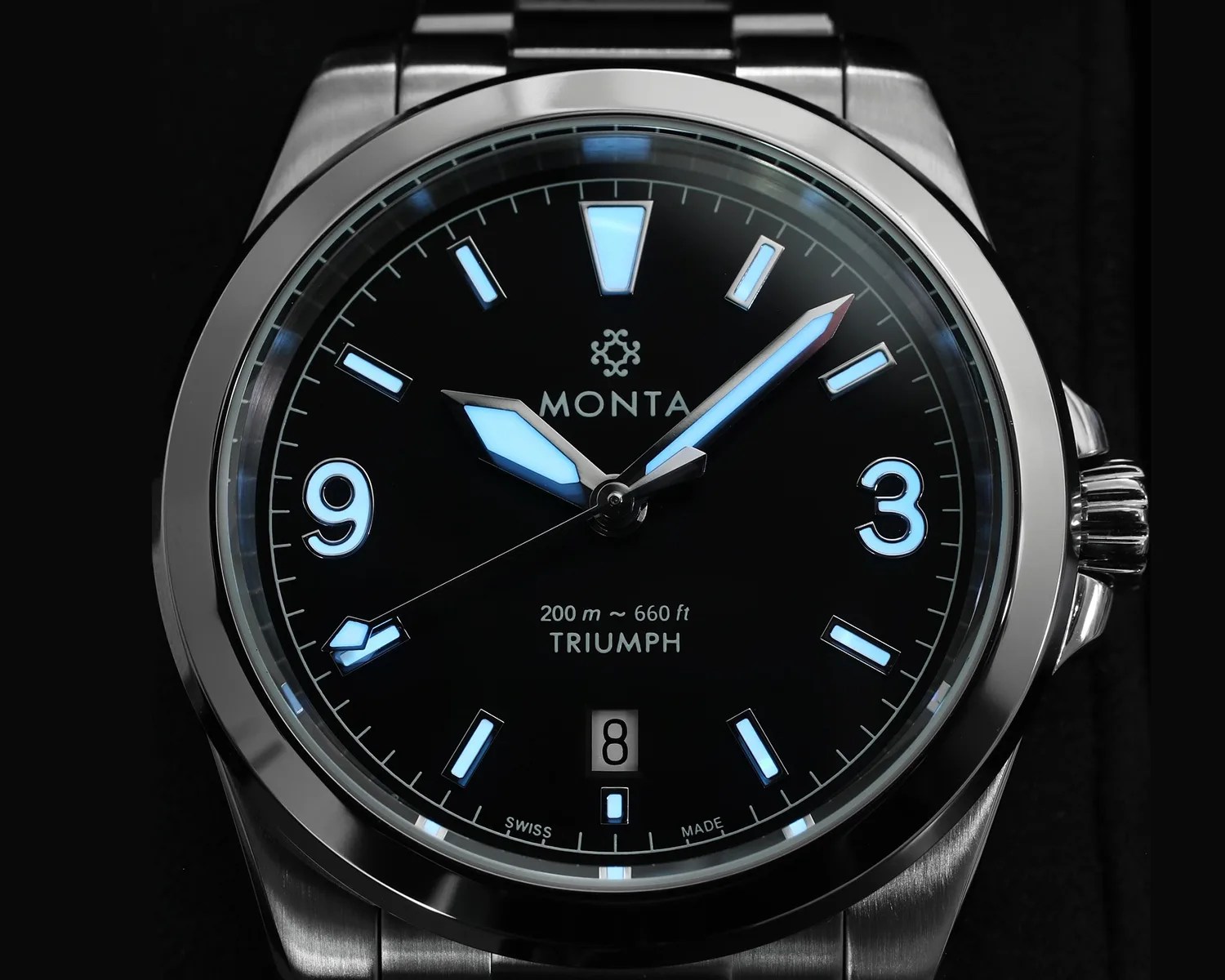 Close-up of a Monta Triumph wristwatch with a black dial, luminescent blue hands and markers, and a stainless steel case and bracelet.