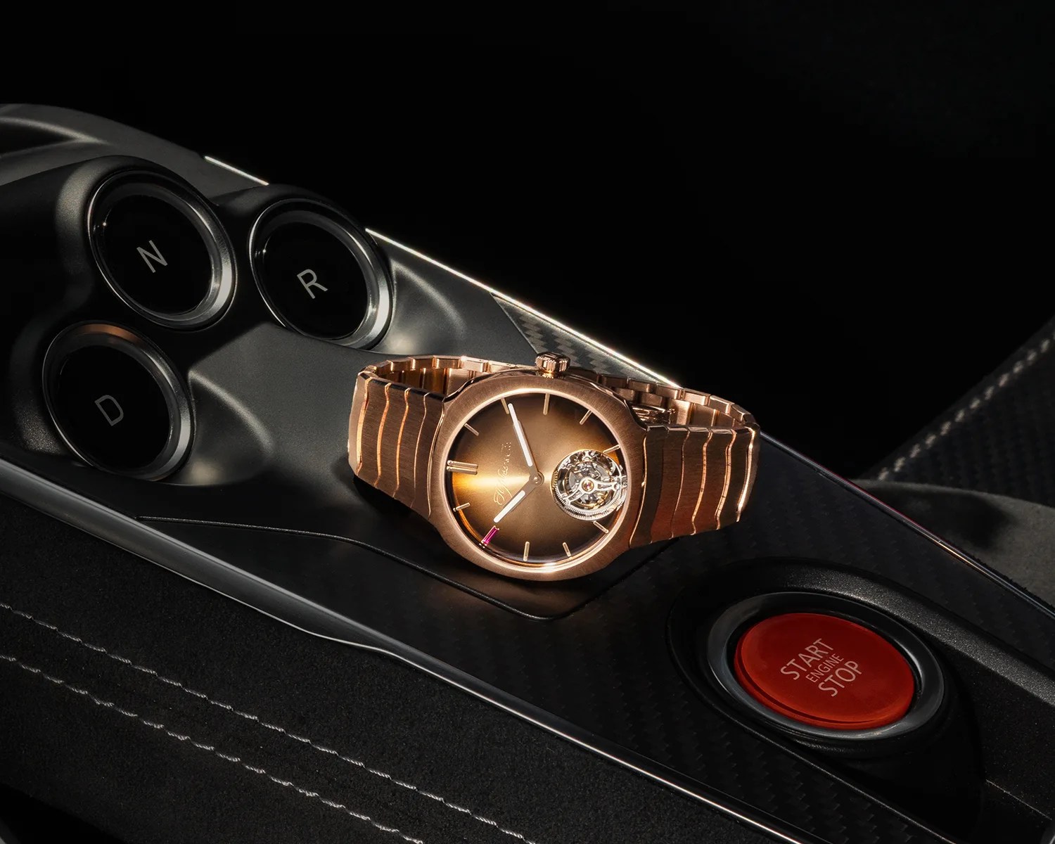 Rose gold wristwatch with a brown dial and visible tourbillon placed on a car's black center console near gear buttons and a red start/stop engine button.