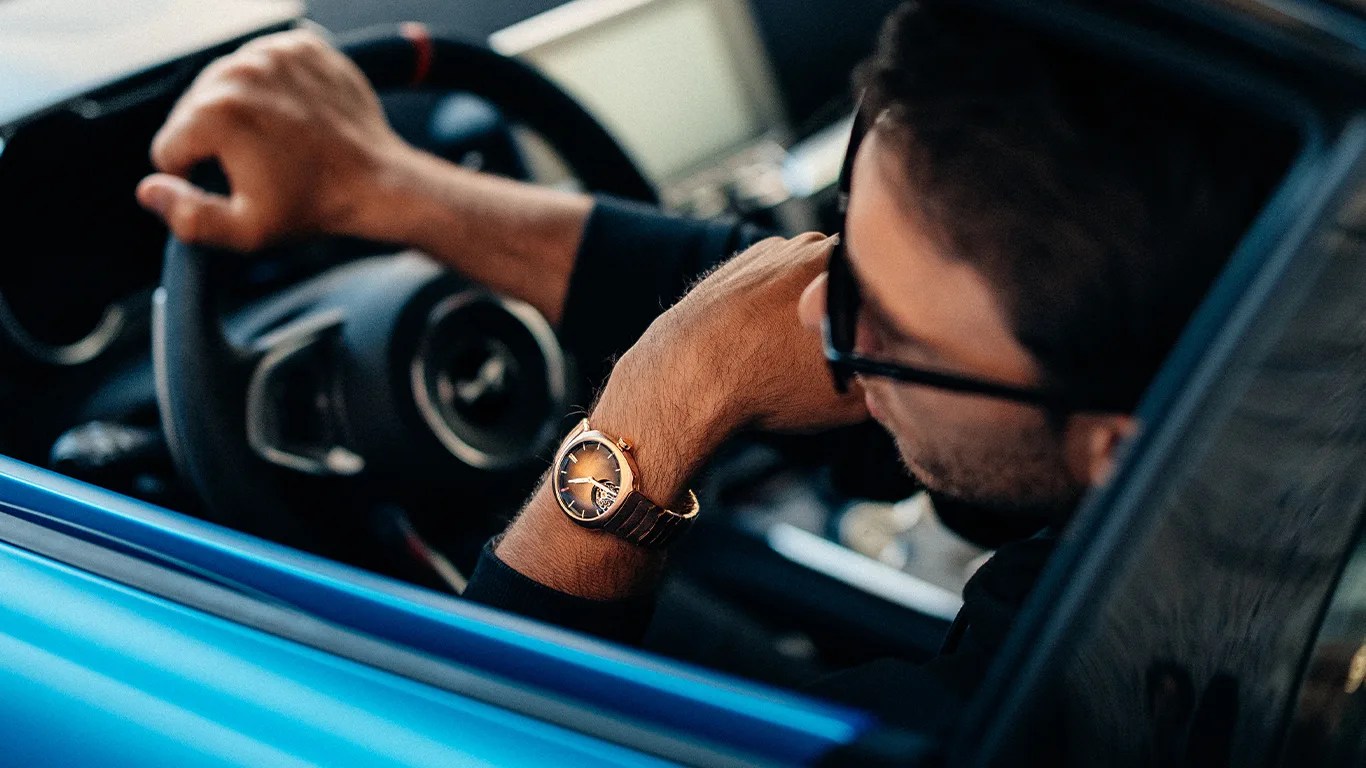 Person wearing a brown leather strap wristwatch with a gold-tone case, resting arm on a blue car door.