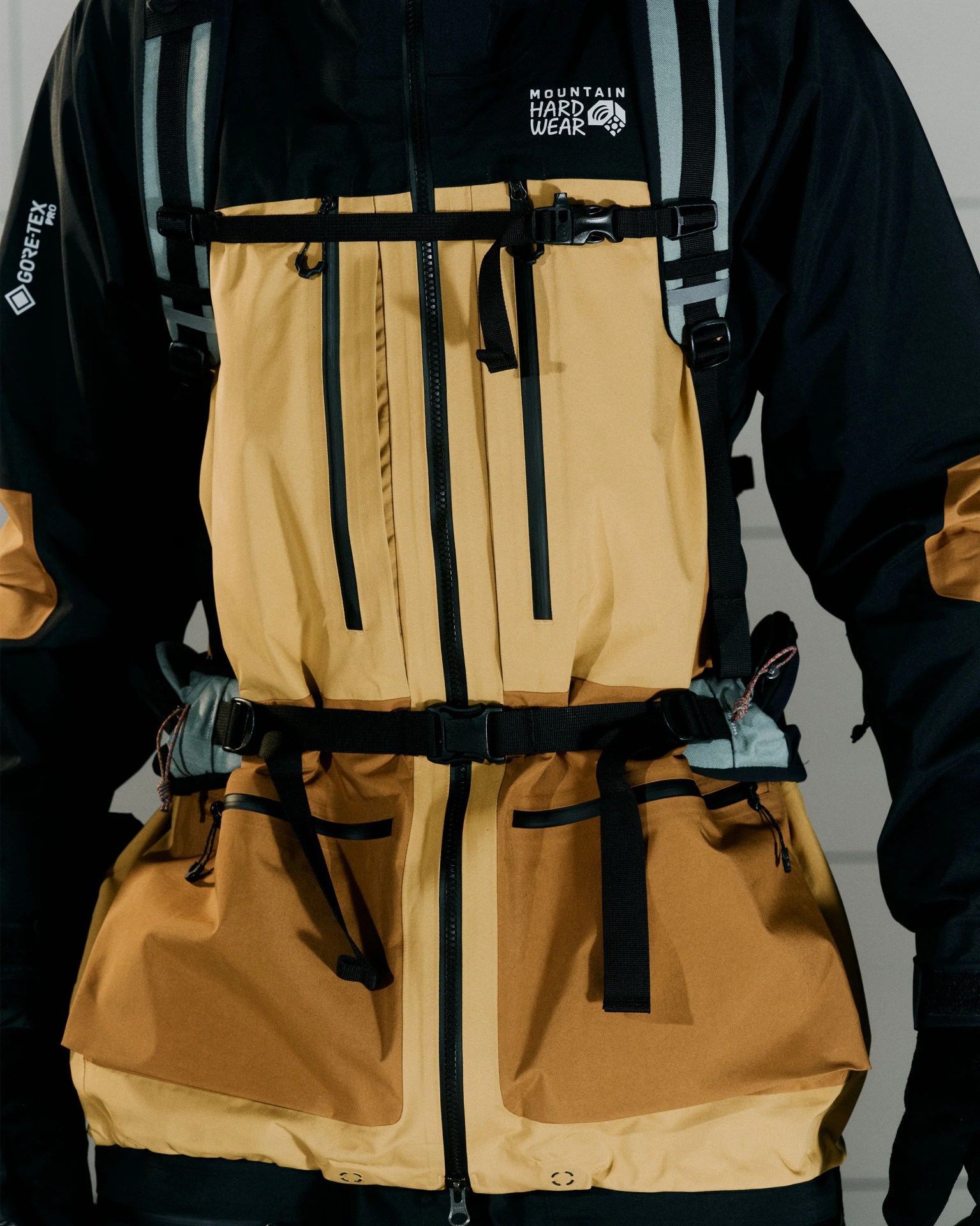 Two-tone tan and black Mountain Hardwear Gore-Tex Pro jacket with multiple black zippers and adjustable straps.