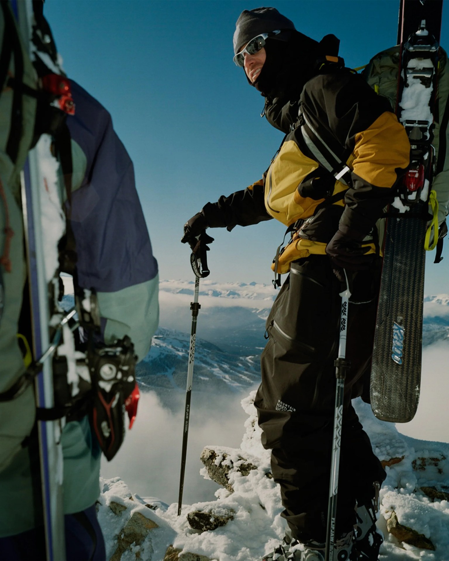Two skiers in winter gear with skis and poles standing on a snowy mountain peak with a clear blue sky.