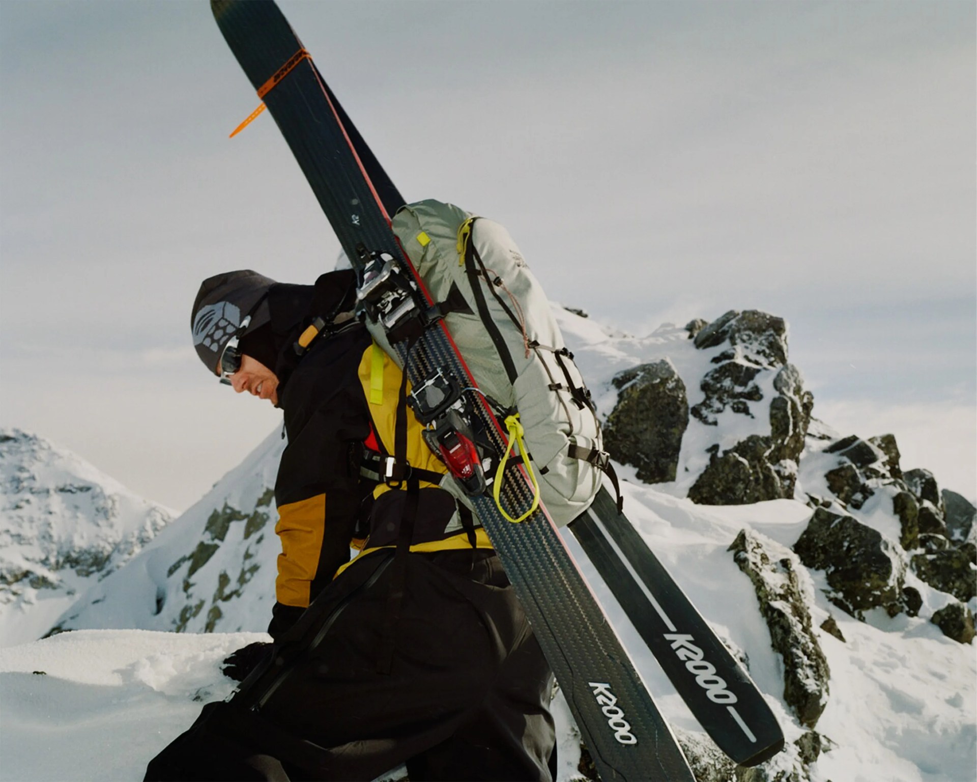 Person wearing a black and yellow jacket with a gray backpack carrying black K2000 skis on a snowy mountain.