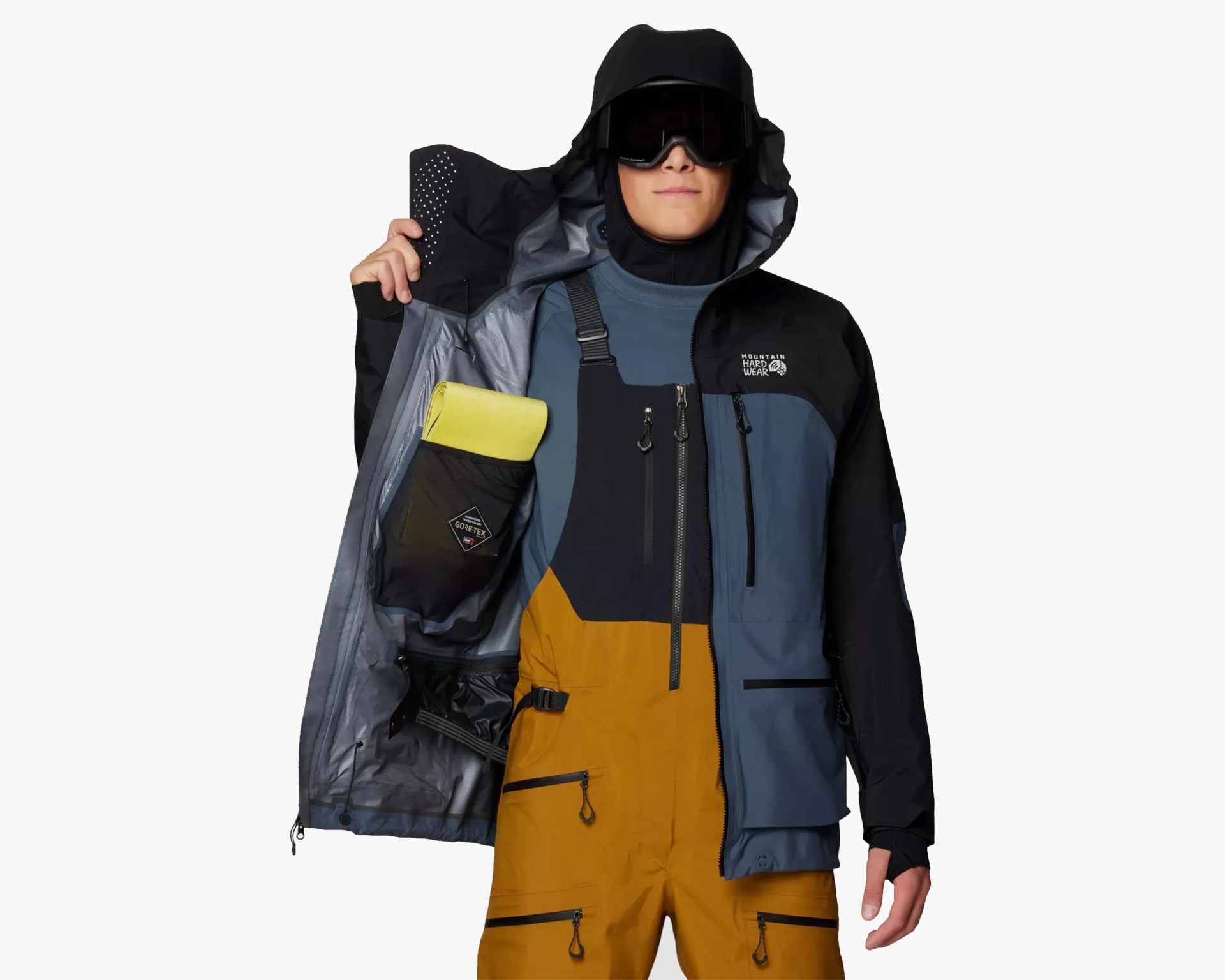 Person wearing a black and blue Mountain Hardwear jacket with hood and goggles, showing the inside pocket holding a yellow item.