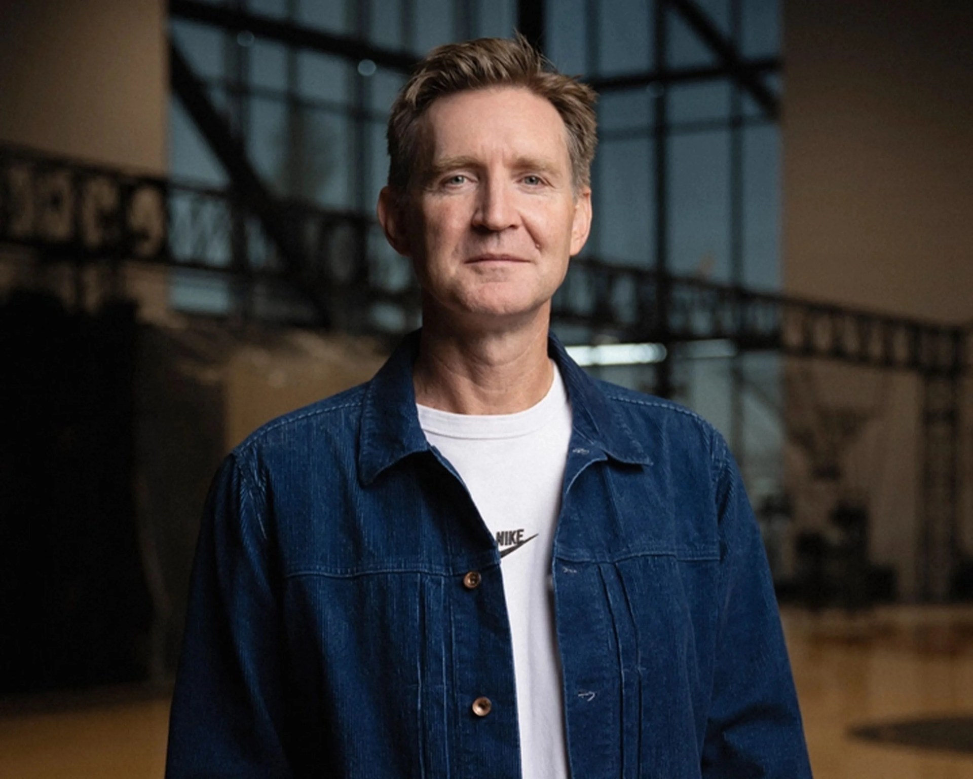Man wearing a blue denim jacket over a white Nike t-shirt standing indoors with blurred background.