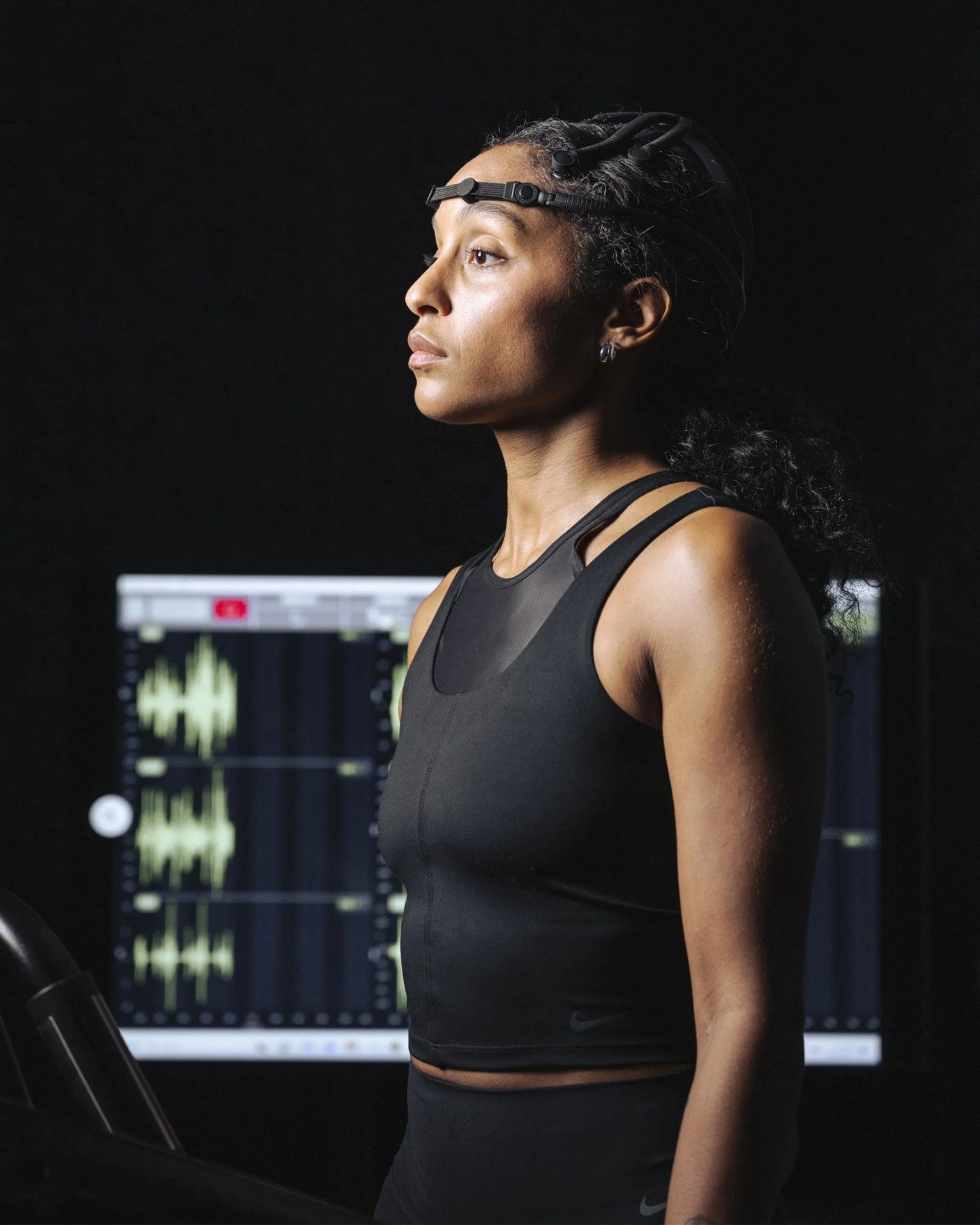 Woman wearing a black sleeveless athletic top and a headband with sensors, standing in front of a screen displaying waveform data.