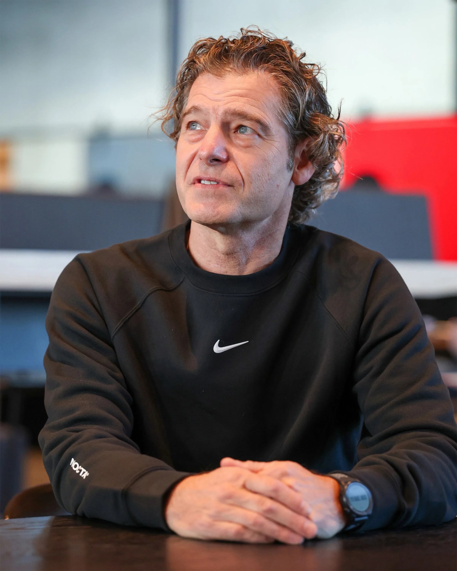 Michael Donaghu wearing a black Nike sweatshirt with a watch, sitting at a table and looking up.