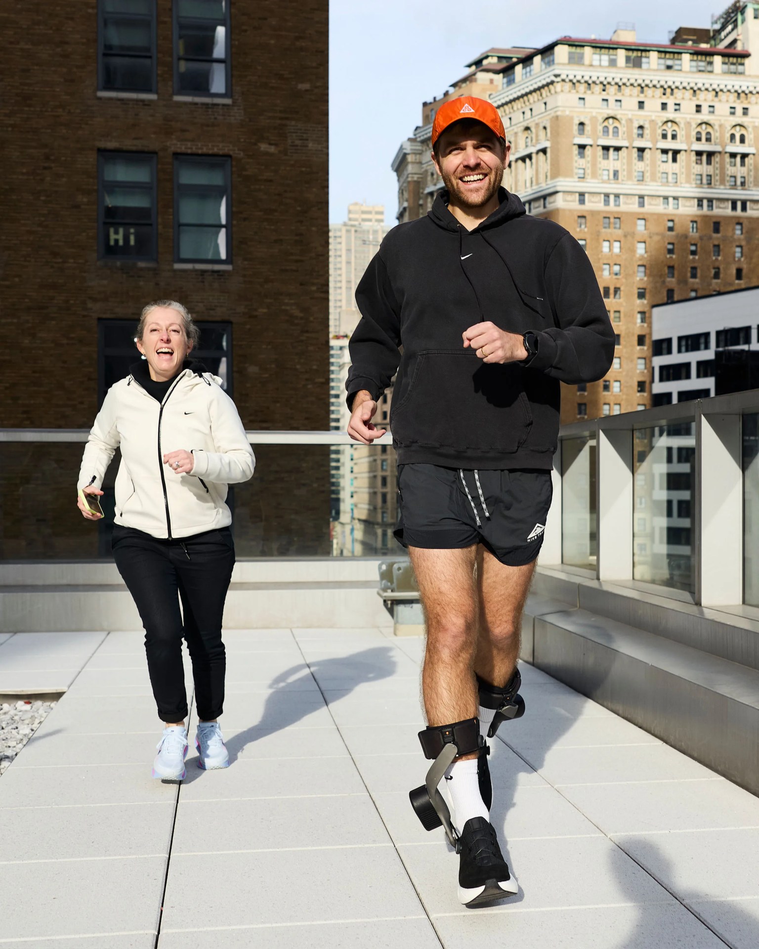 Two people jogging on a rooftop terrace, one wearing black shorts and ankle braces, the other in black pants and a white jacket.