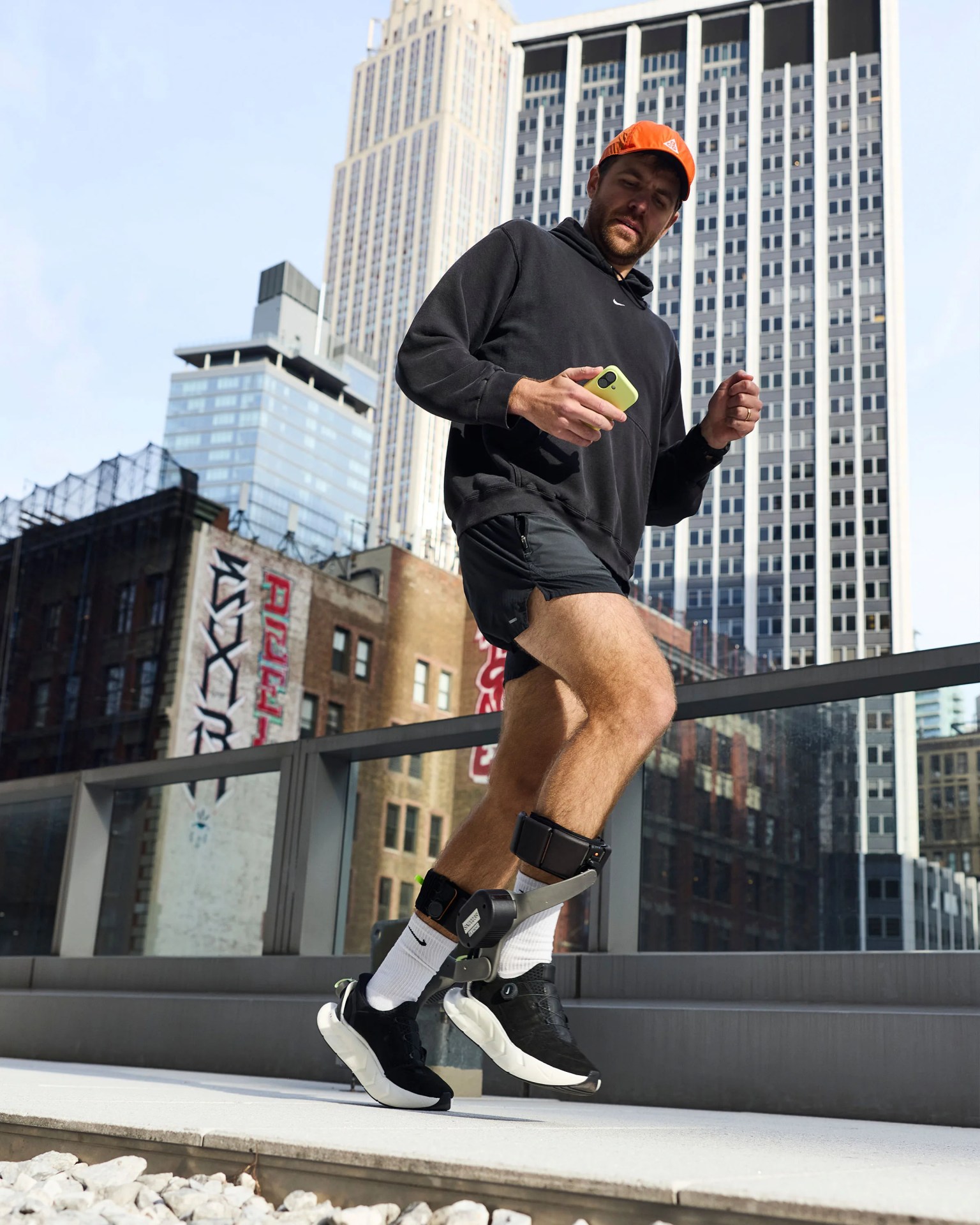 Man wearing black shorts, black hoodie, and orange cap using a leg-worn fitness device while holding a green smartphone in an urban setting.