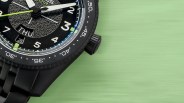 Close-up of a black wristwatch with white numerals, a textured dial, day and date display, and a black metal band on a green surface.