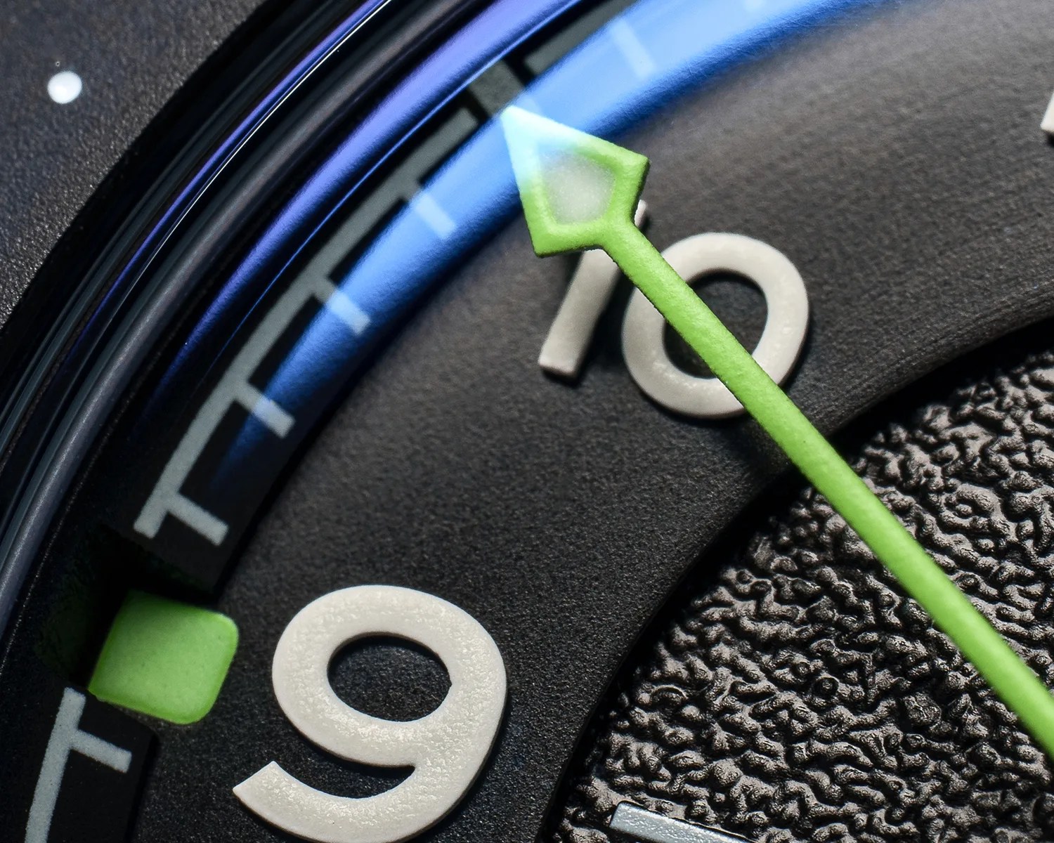Close-up of a black watch face showing white numbers 9 and 10, with a green arrow-shaped hand and textured dial.
