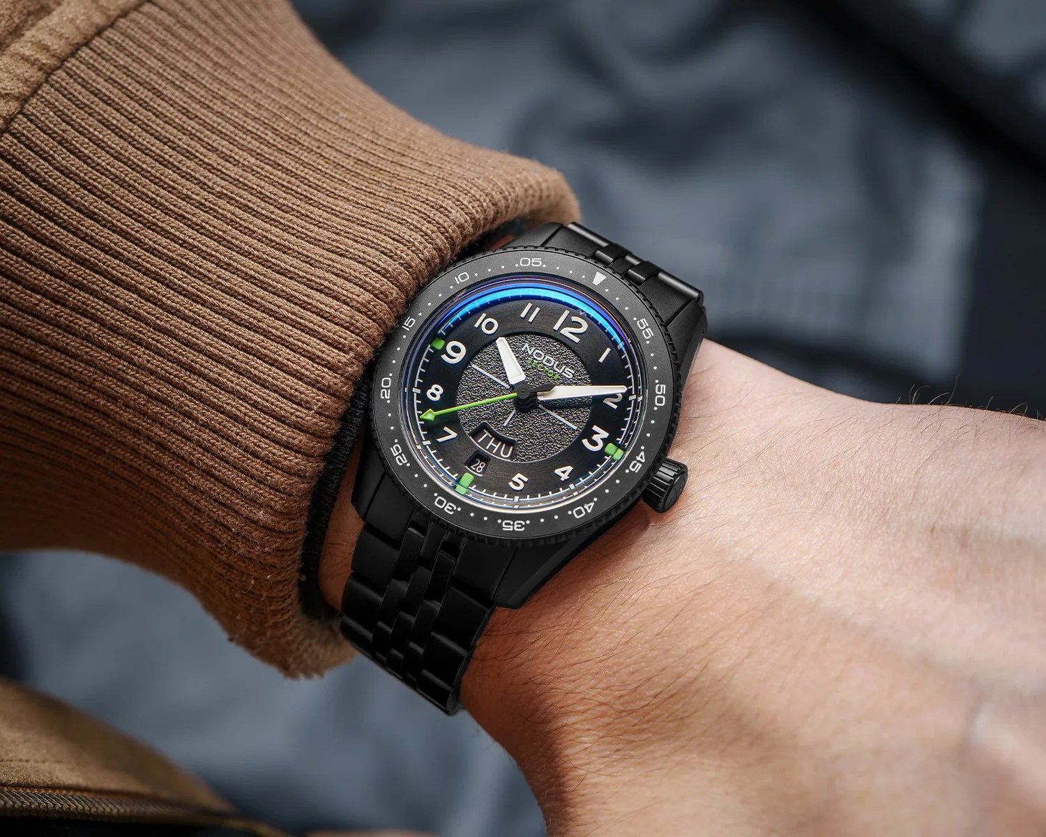 Black metal wristwatch with textured black dial, white numerals, green accents, and day-date display worn on a wrist.