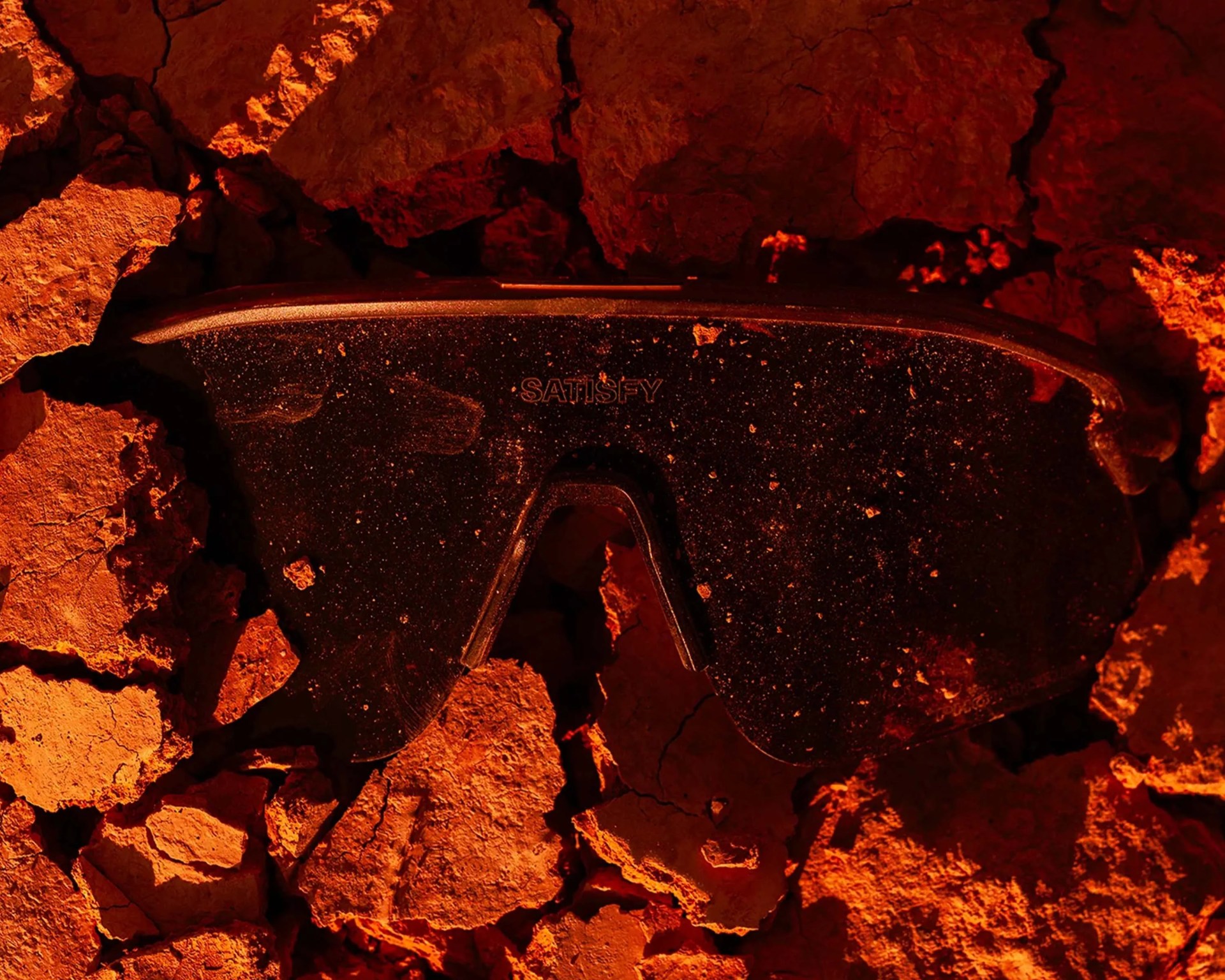 Black Satisfy brand sports sunglasses covered in dust on cracked dry earth.