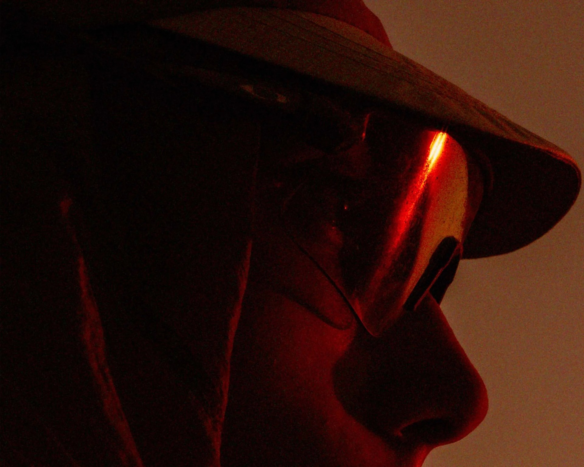Close-up of a person wearing reflective sunglasses and a wide-brimmed hat, illuminated by red light.