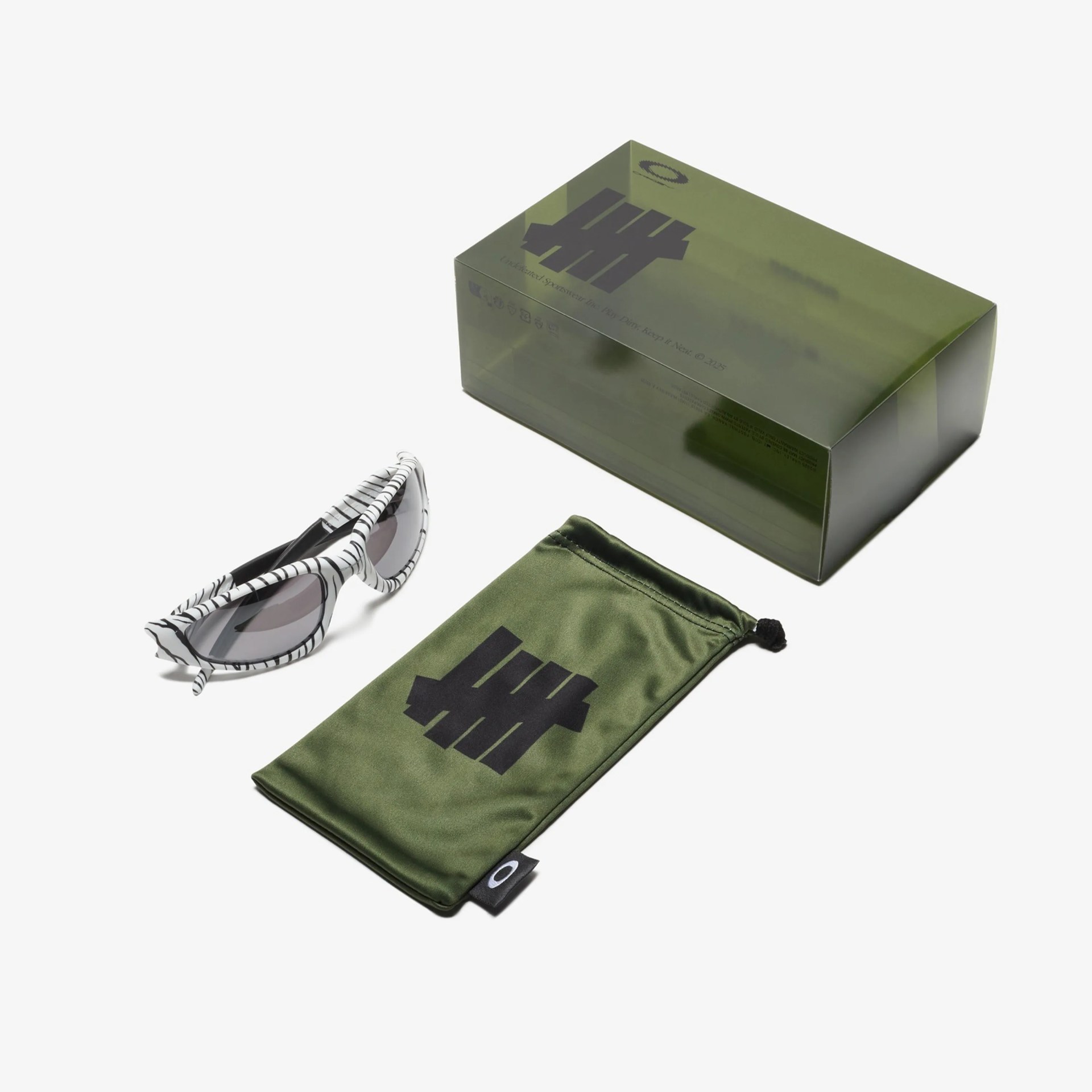 White and black zebra-striped sunglasses with gray lenses next to an olive green drawstring pouch and matching box.