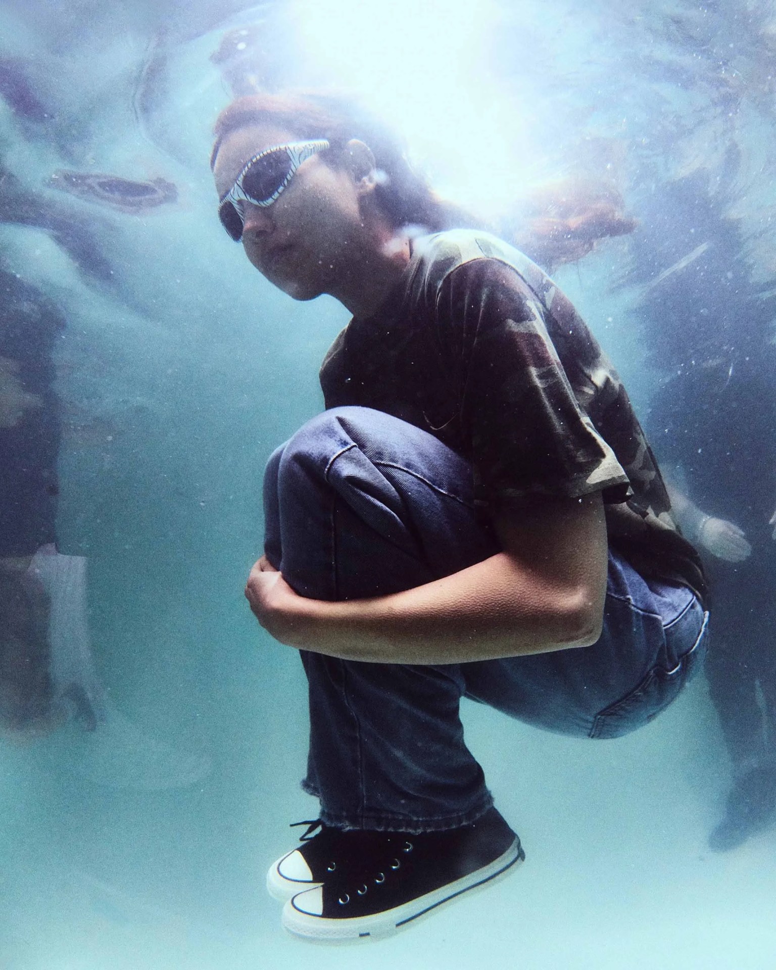 Person wearing zebra-striped sunglasses, camouflage shirt, blue jeans, and black sneakers crouching underwater.