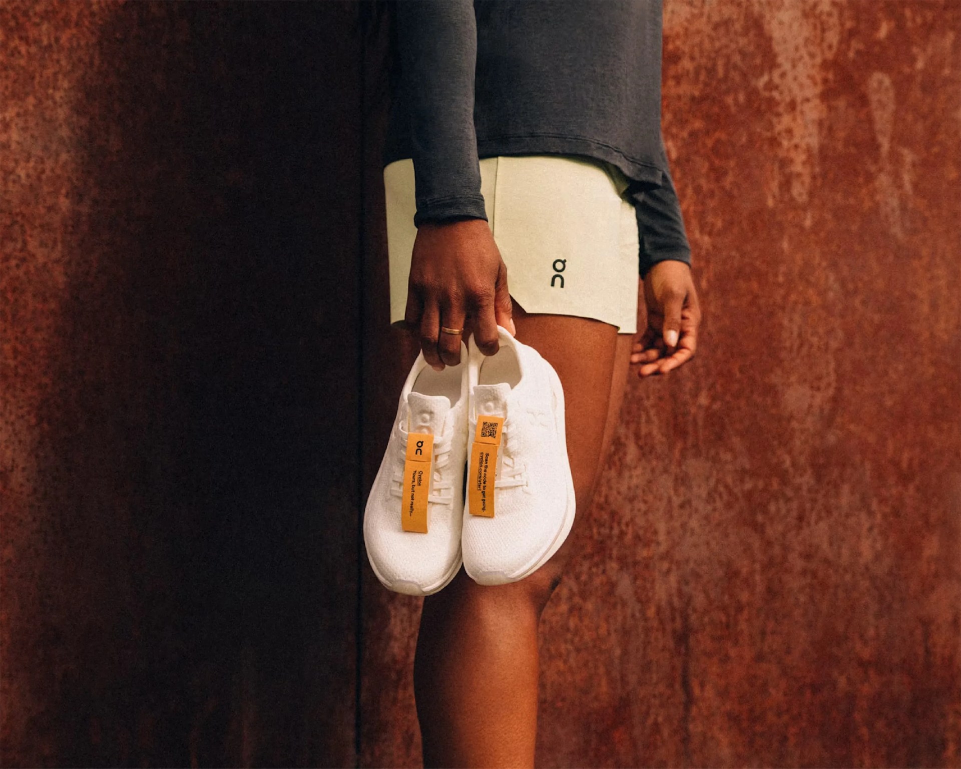Person holding white running shoes with orange tags, wearing light beige shorts with "on" logo and a dark long-sleeve top.