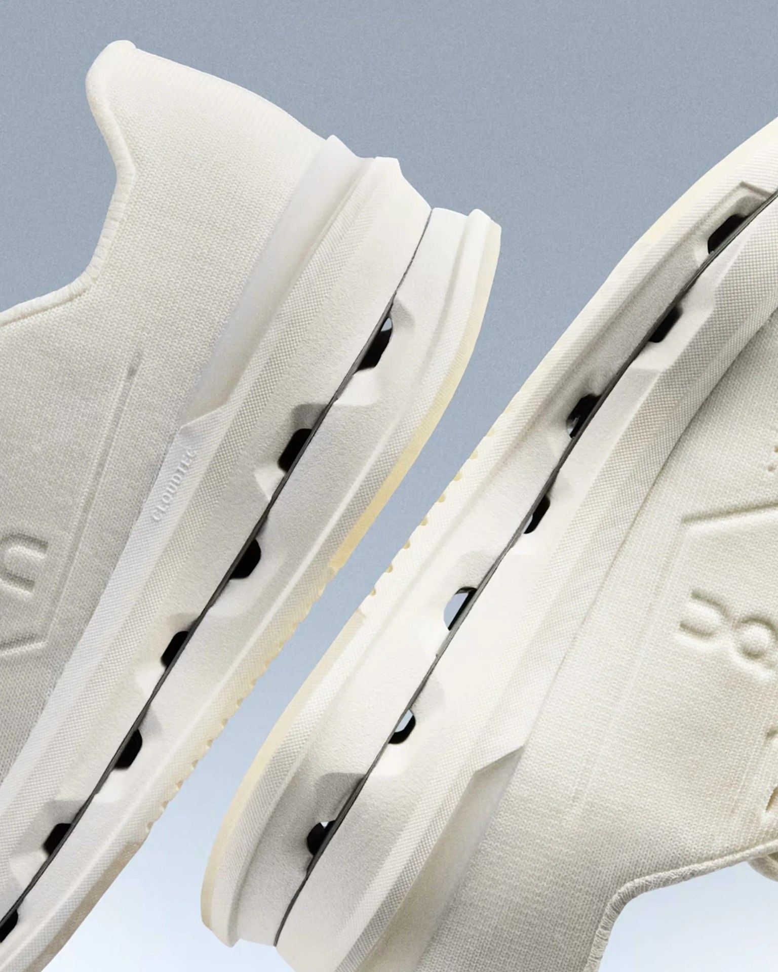 Close-up of two white athletic shoes with textured fabric uppers and thick soles featuring black circular cutouts.