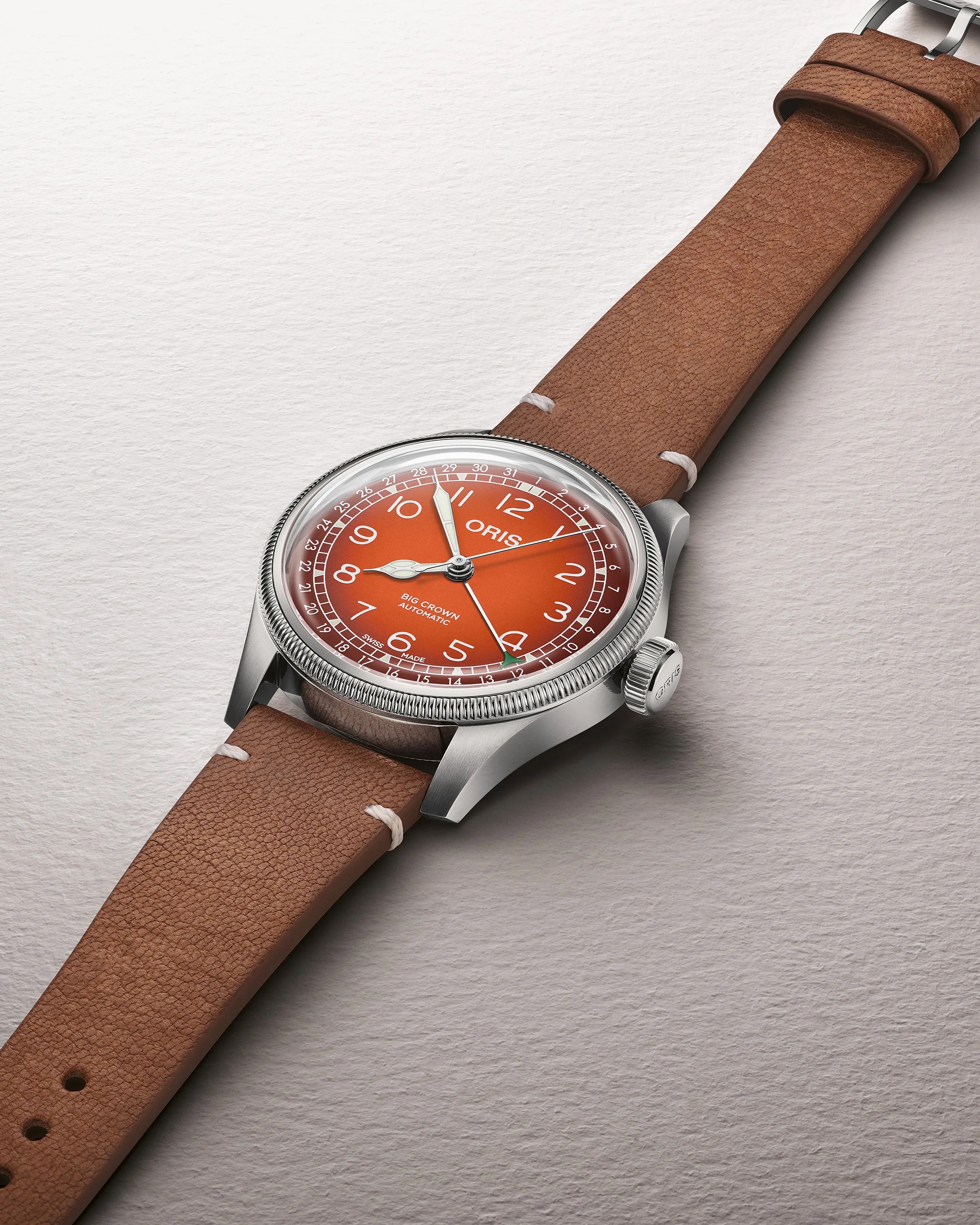 Stainless steel Oris Big Crown automatic watch with orange dial and brown leather strap on a textured white surface.