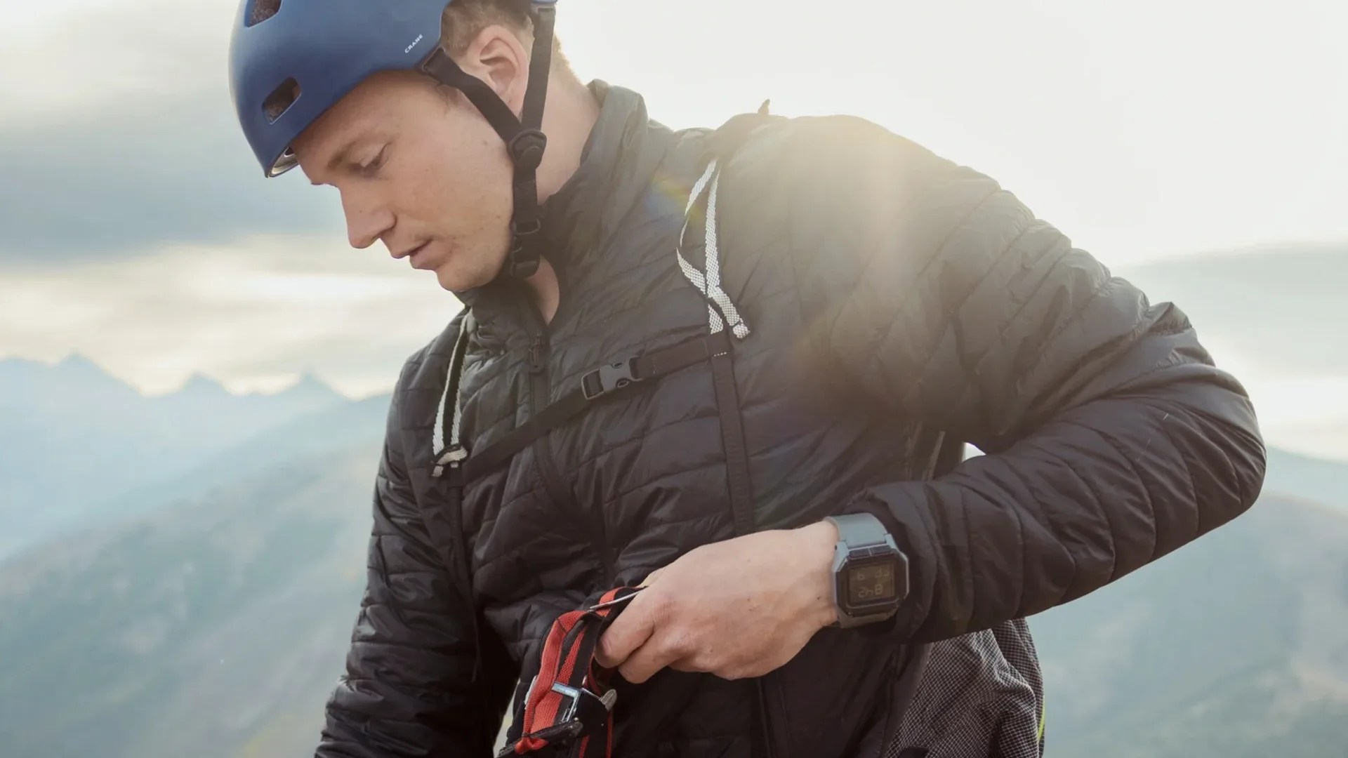 Person wearing a blue helmet, black quilted jacket, and digital watch adjusting red climbing gear outdoors with mountains in background.