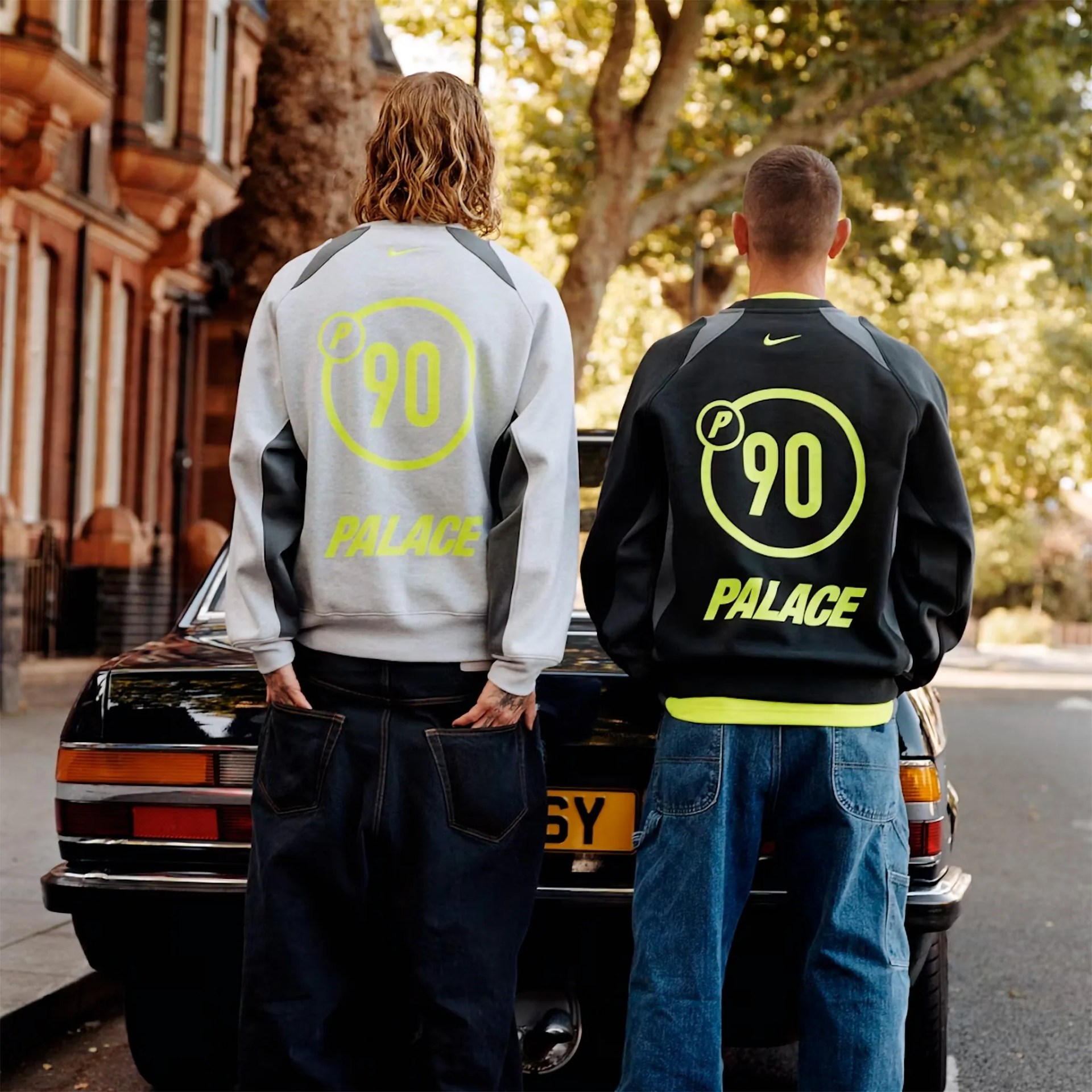 Two people wearing Palace P-90 sweatshirts, one gray and one black, standing in front of a black car on a street.