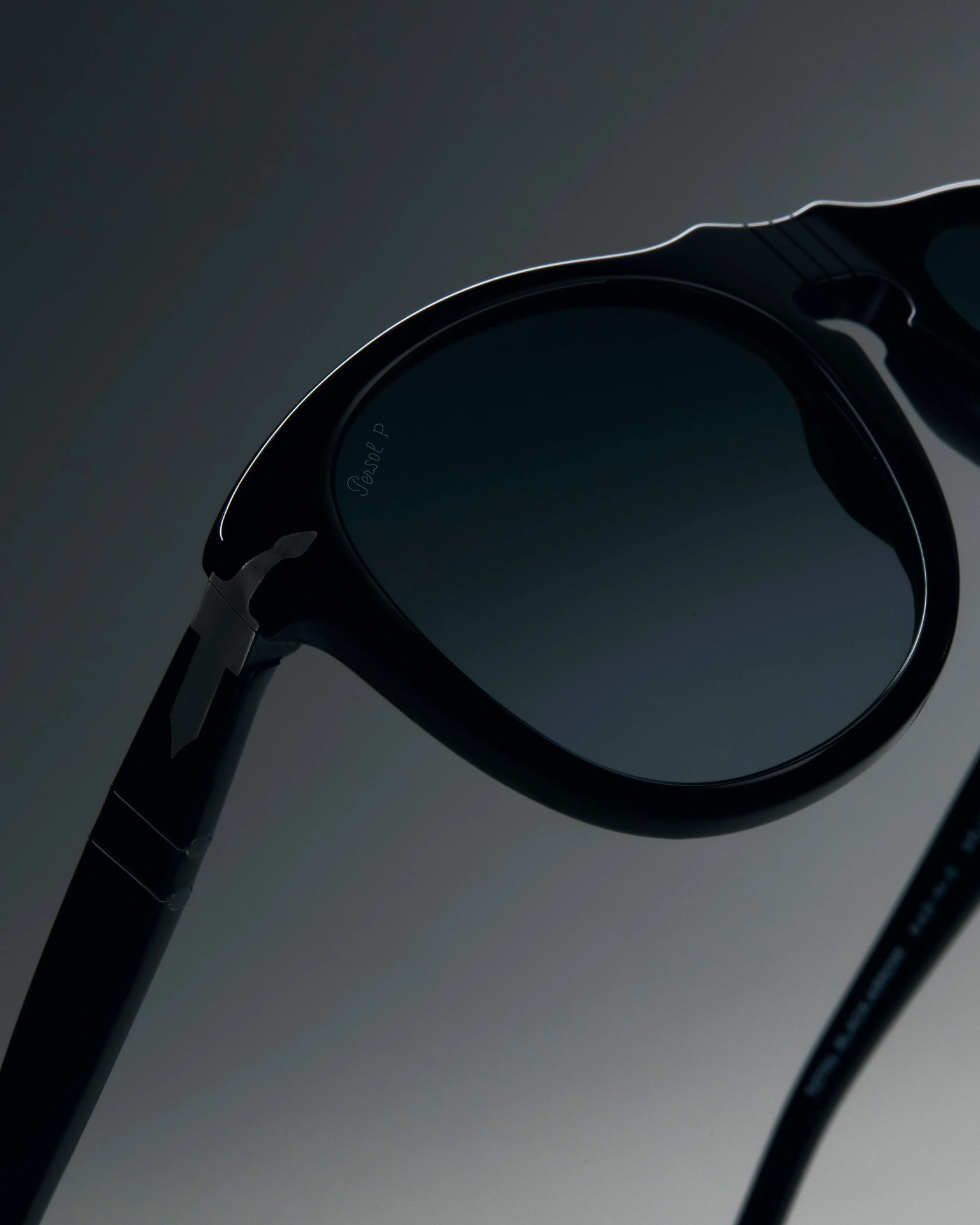 Close-up of black sunglasses with dark tinted lenses and a subtle logo on the temple against a dark gradient background.