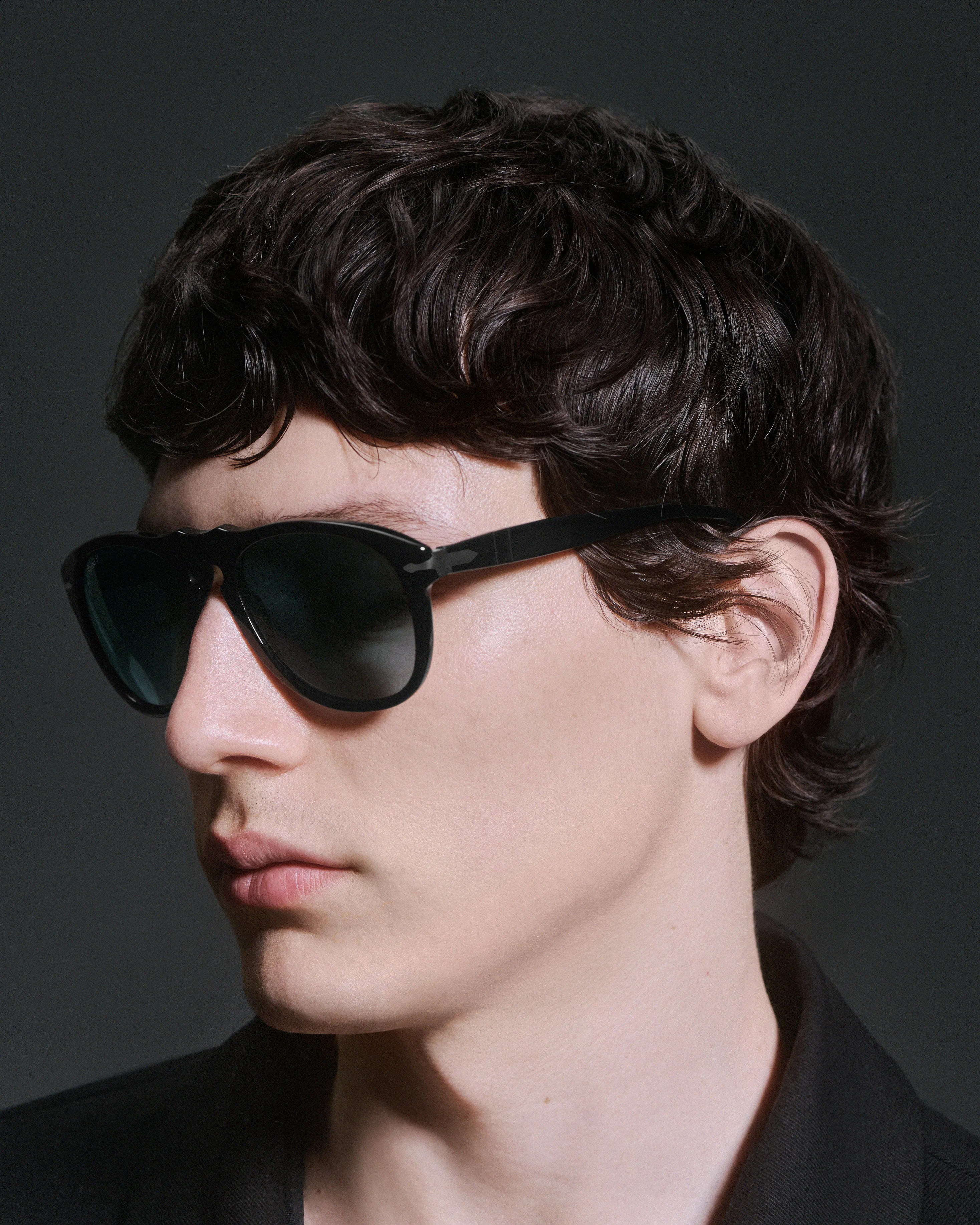 Close-up of a person wearing black rectangular sunglasses with dark lenses and a black jacket against a dark background.