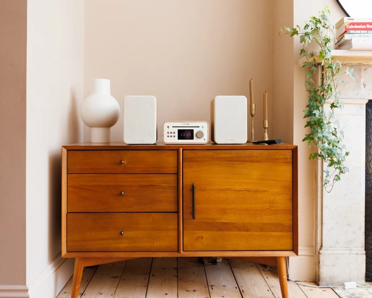 Mid-century wooden sideboard with white speakers, a white radio, a white vase, and two brass candlesticks on top.
