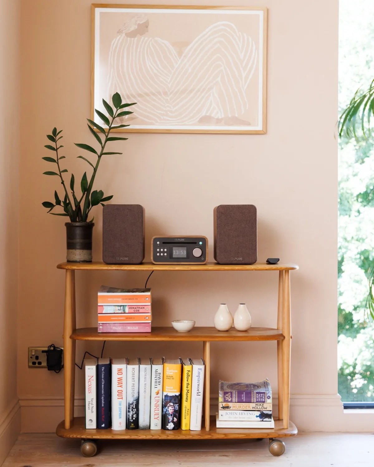 Wooden three-tier shelf with books, a potted plant, a radio with two speakers, and small ceramic vases against a beige wall.