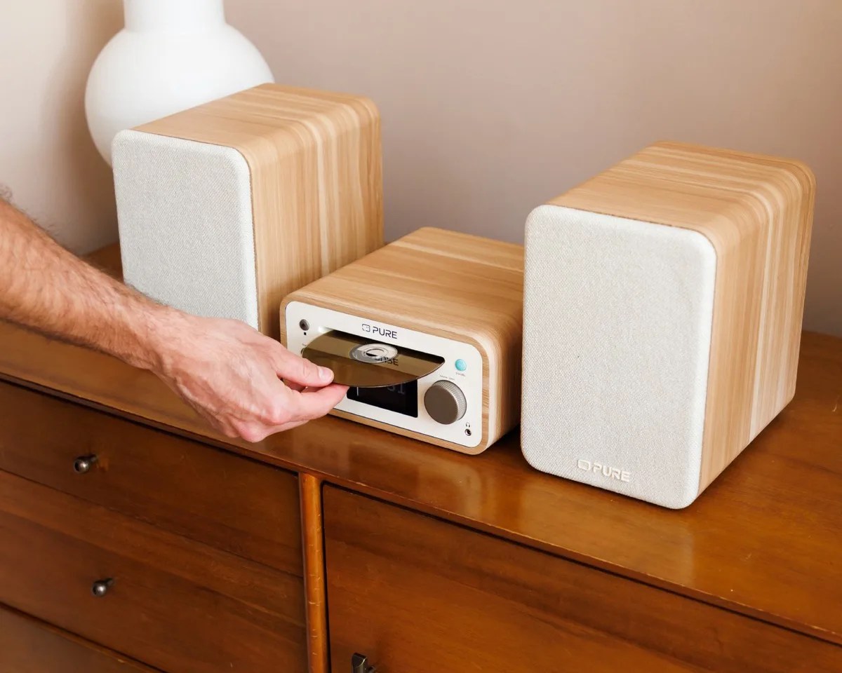 Compact wooden stereo system with two speakers and a person inserting a CD into the central unit on a wooden dresser.