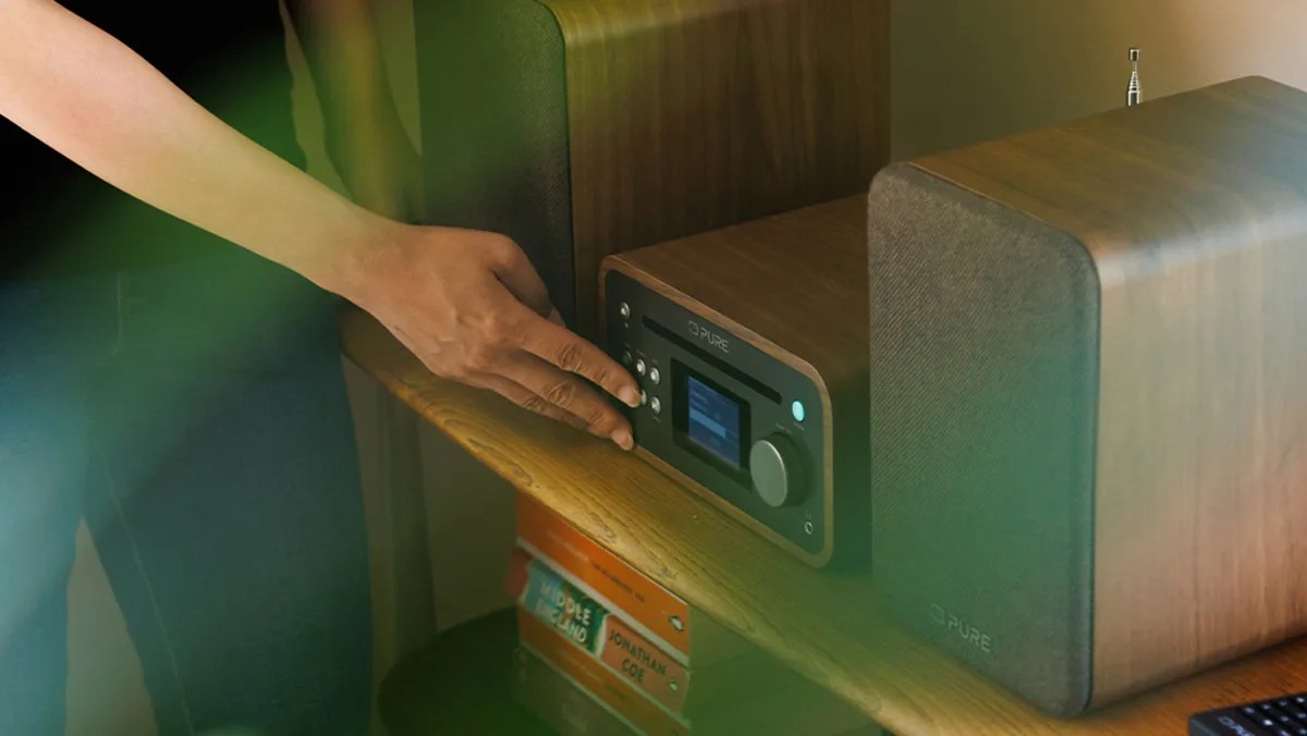 Person adjusting buttons on a Pure brand wooden tabletop radio with two speakers on a wooden shelf.