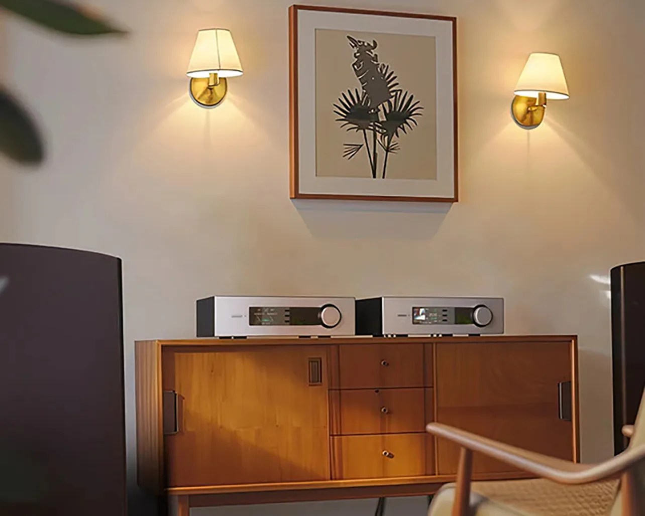 Wooden sideboard with two silver audio components on top, flanked by wall sconces and a framed abstract plant artwork above.