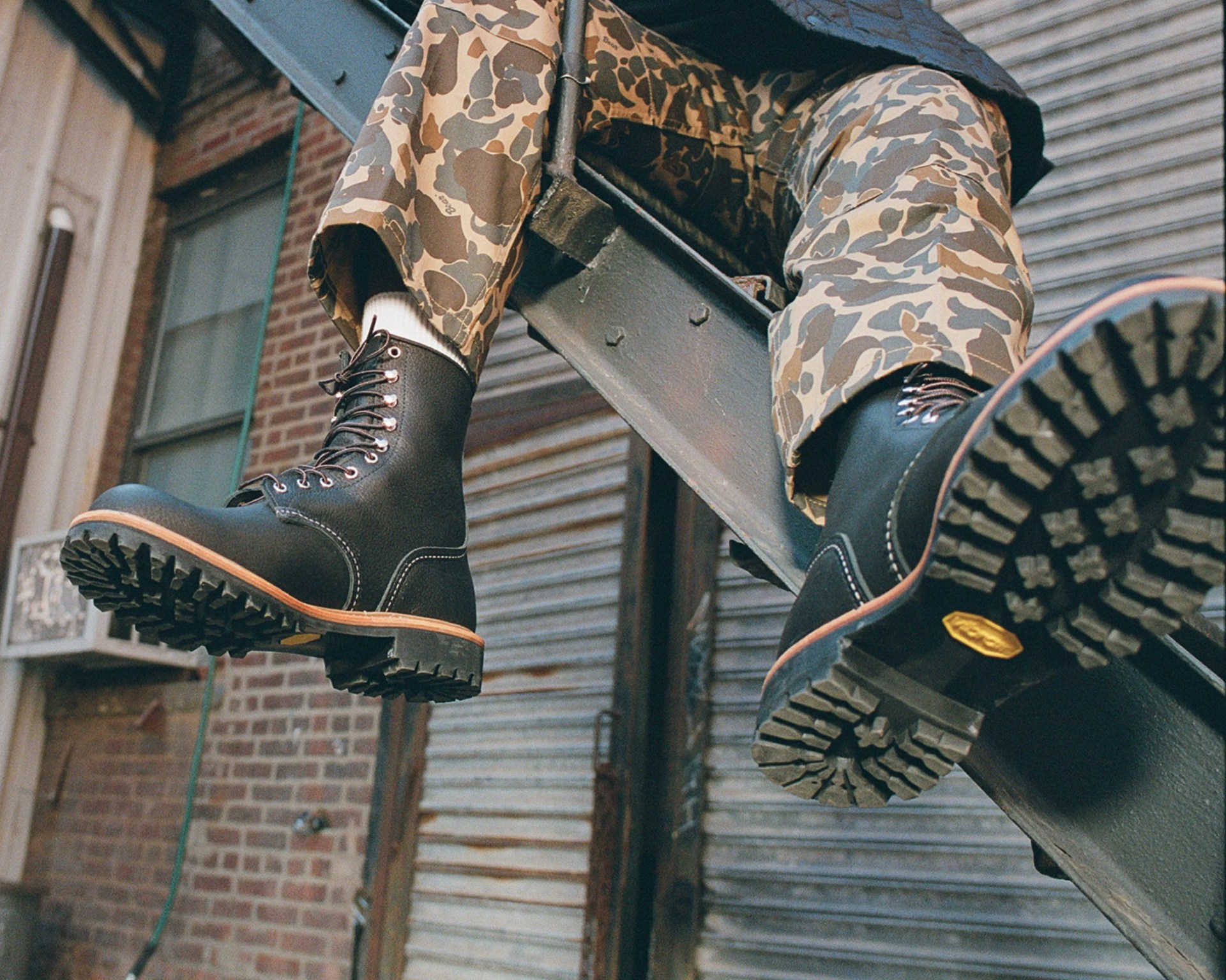 Black leather lace-up boots with thick lug soles worn with camouflage pants, viewed from below.